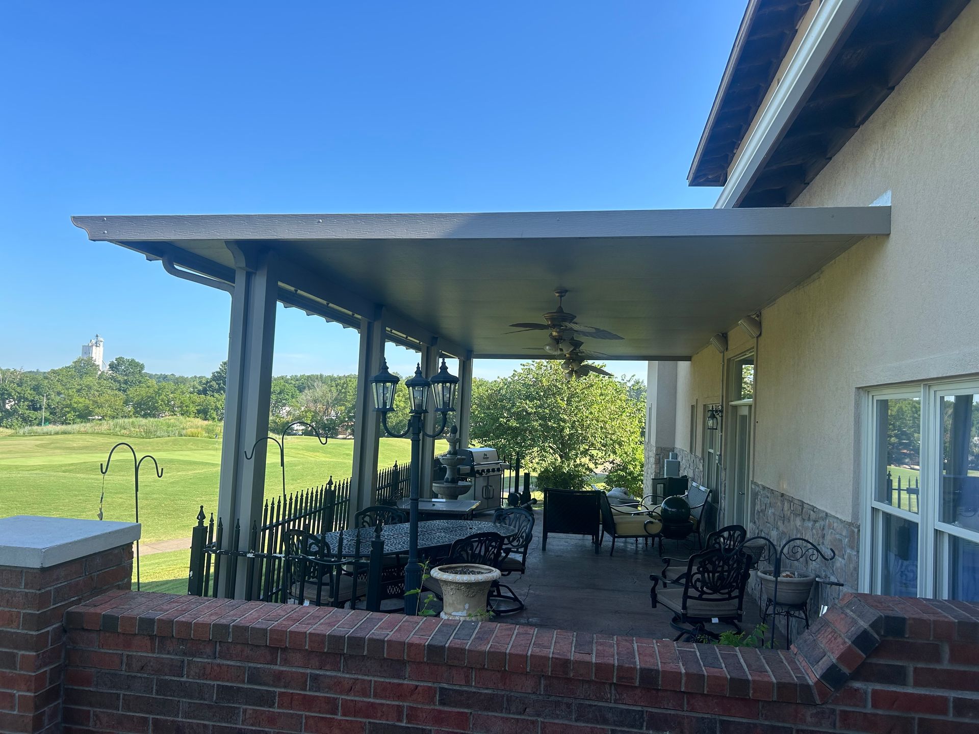 Covered patio with outdoor seating beside a house, overlooking a green field under a blue sky