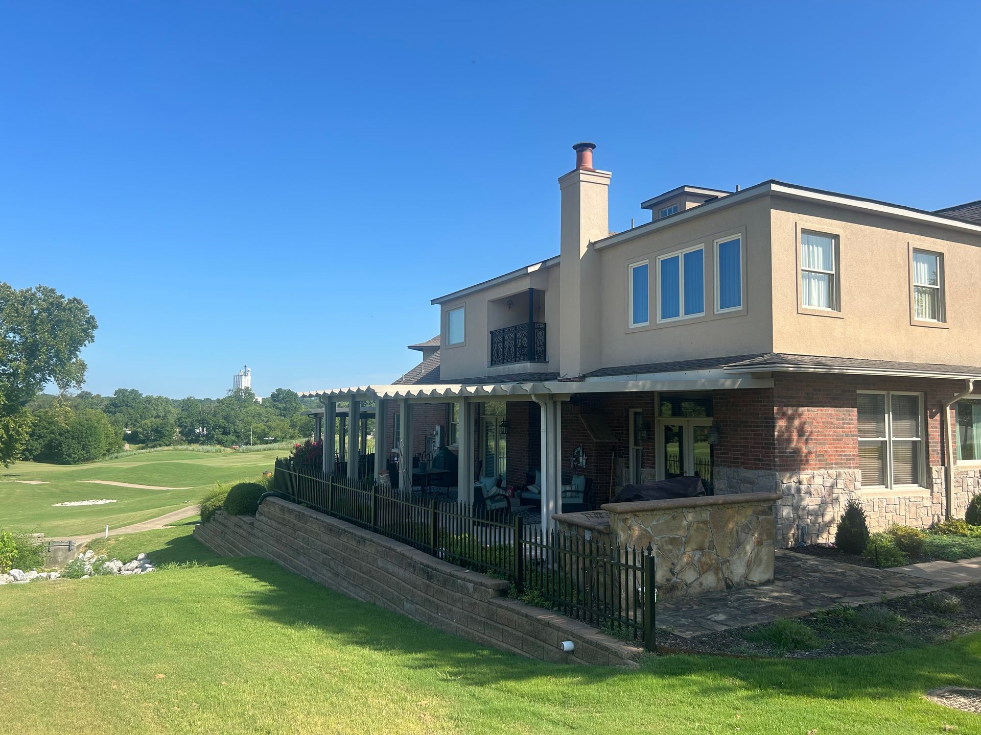 Large beige house with a covered porch beside a green lawn and golf course under a clear blue sky