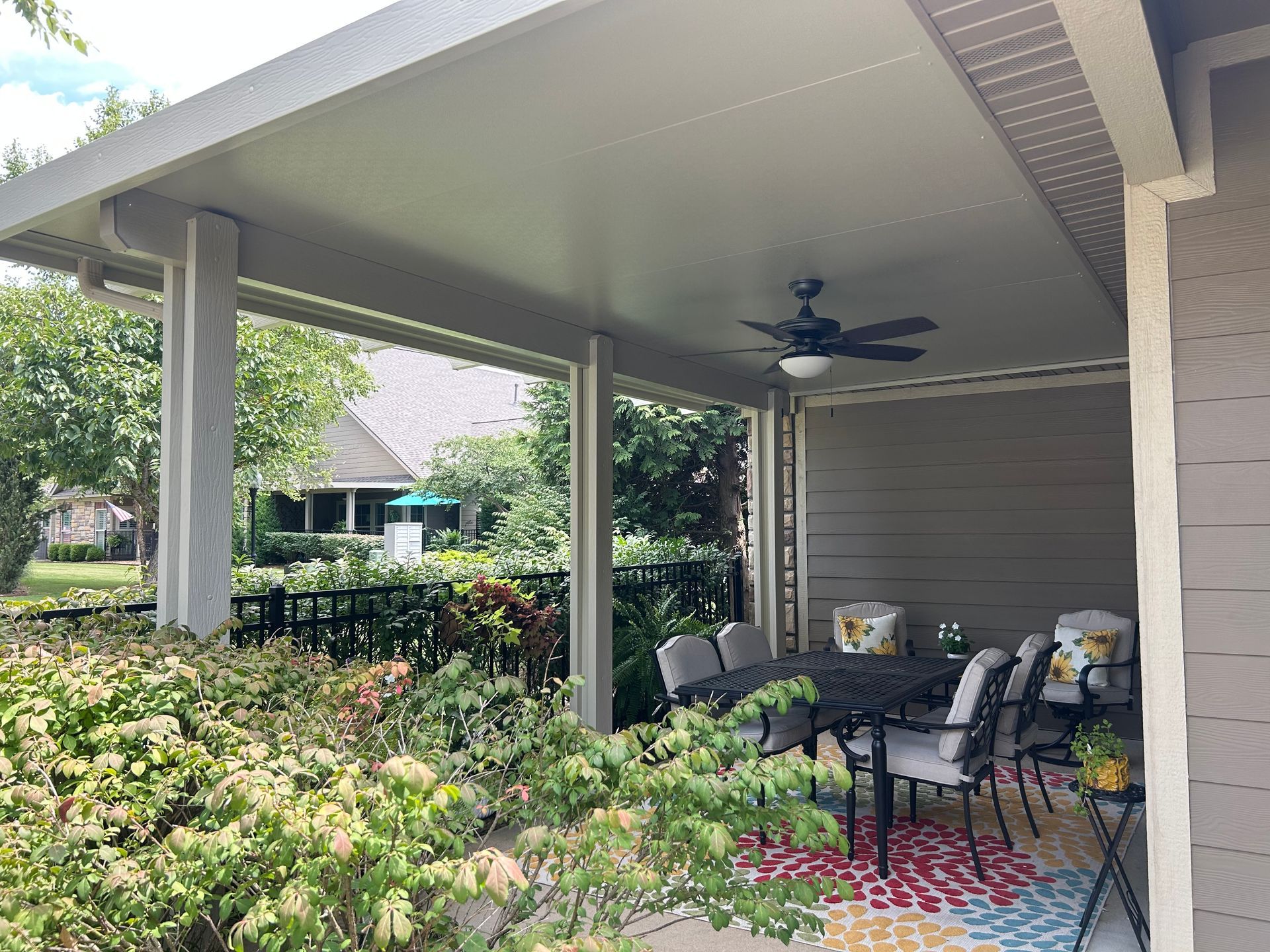 Covered patio with dining table, chairs, ceiling fan, and potted plants beside a house