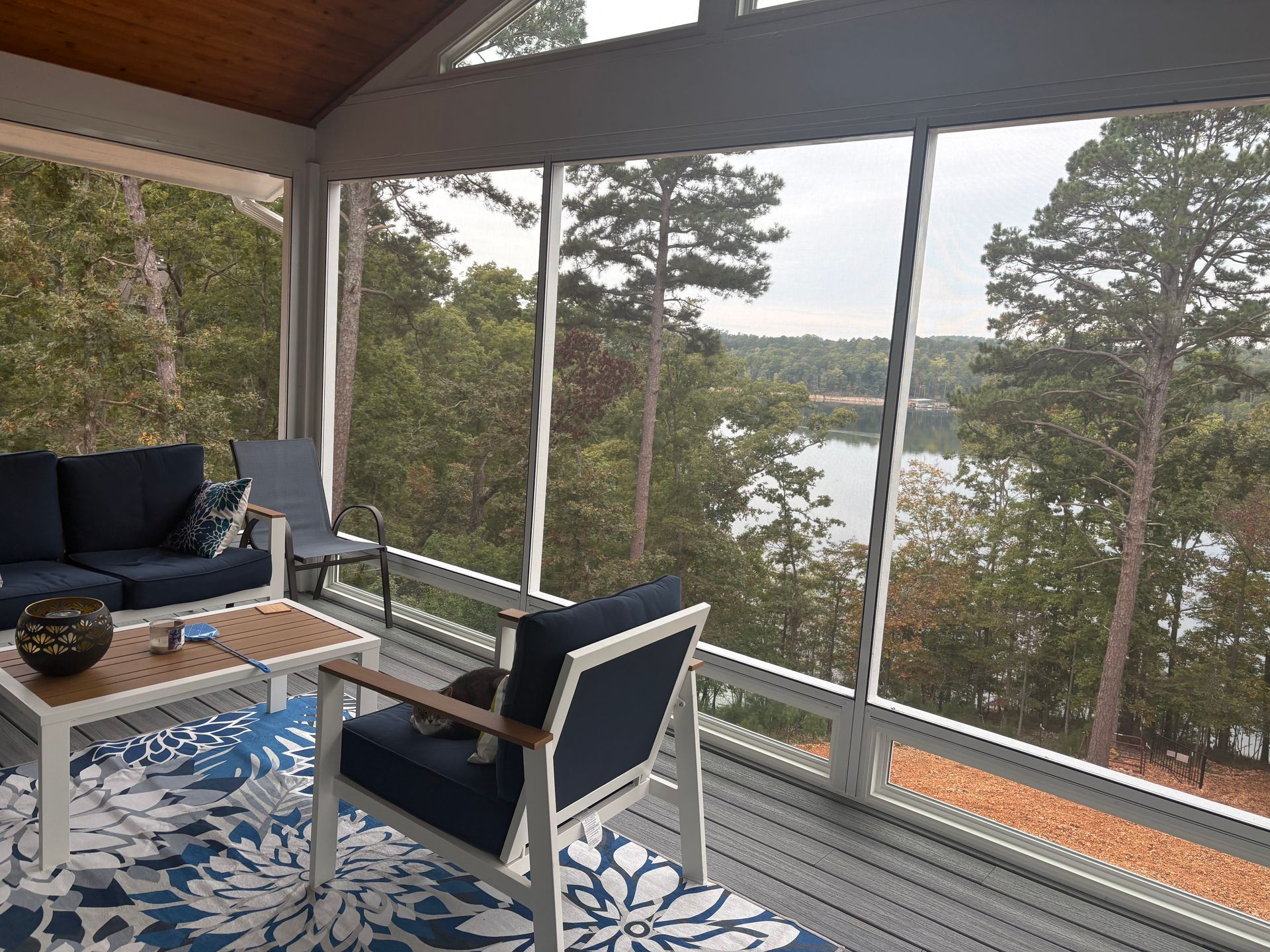 Covered porch with blue chairs and a table overlooking a lake through tall windows.