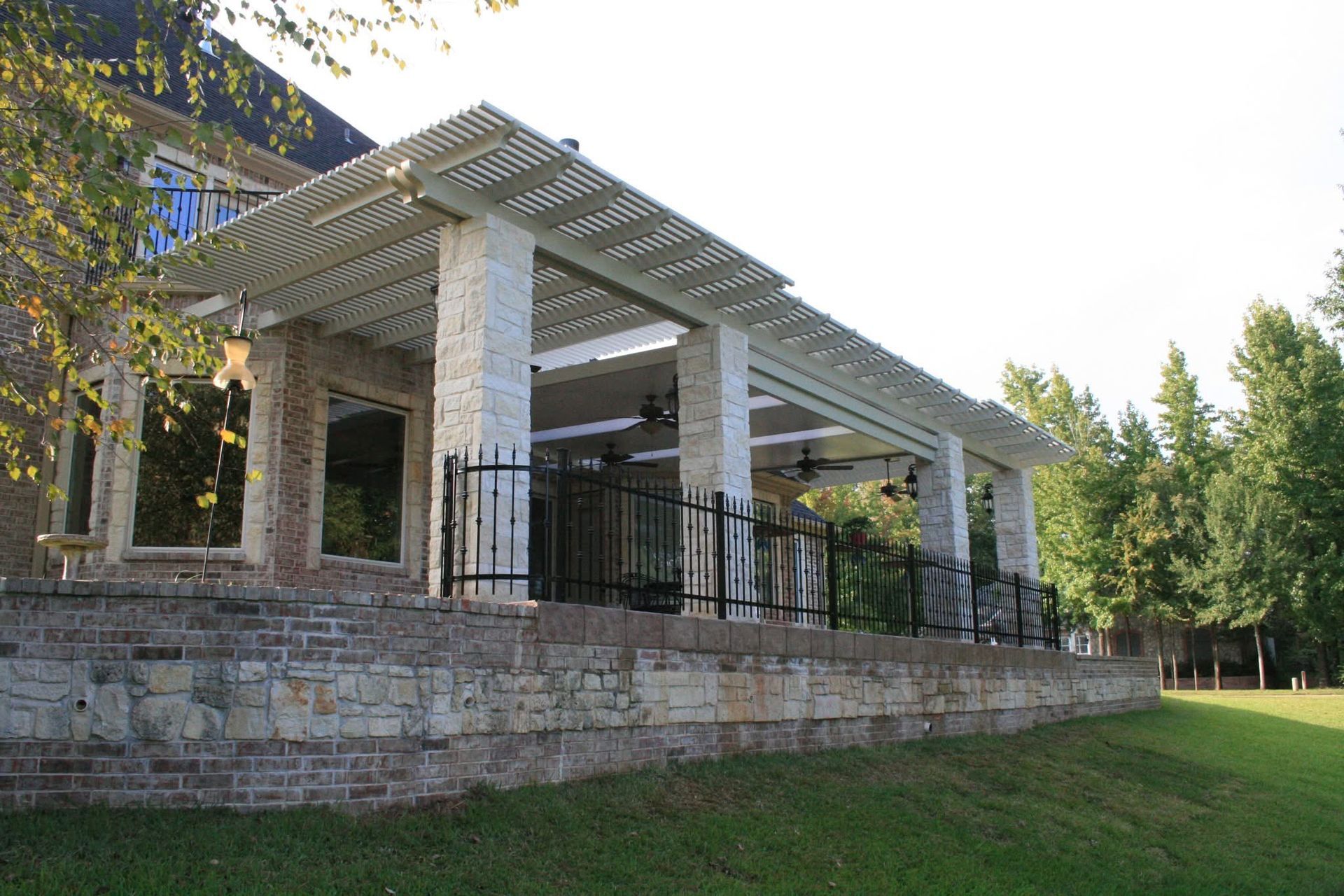 Stone pavilion with white columns and pergola roof on a grassy lawn beside trees
