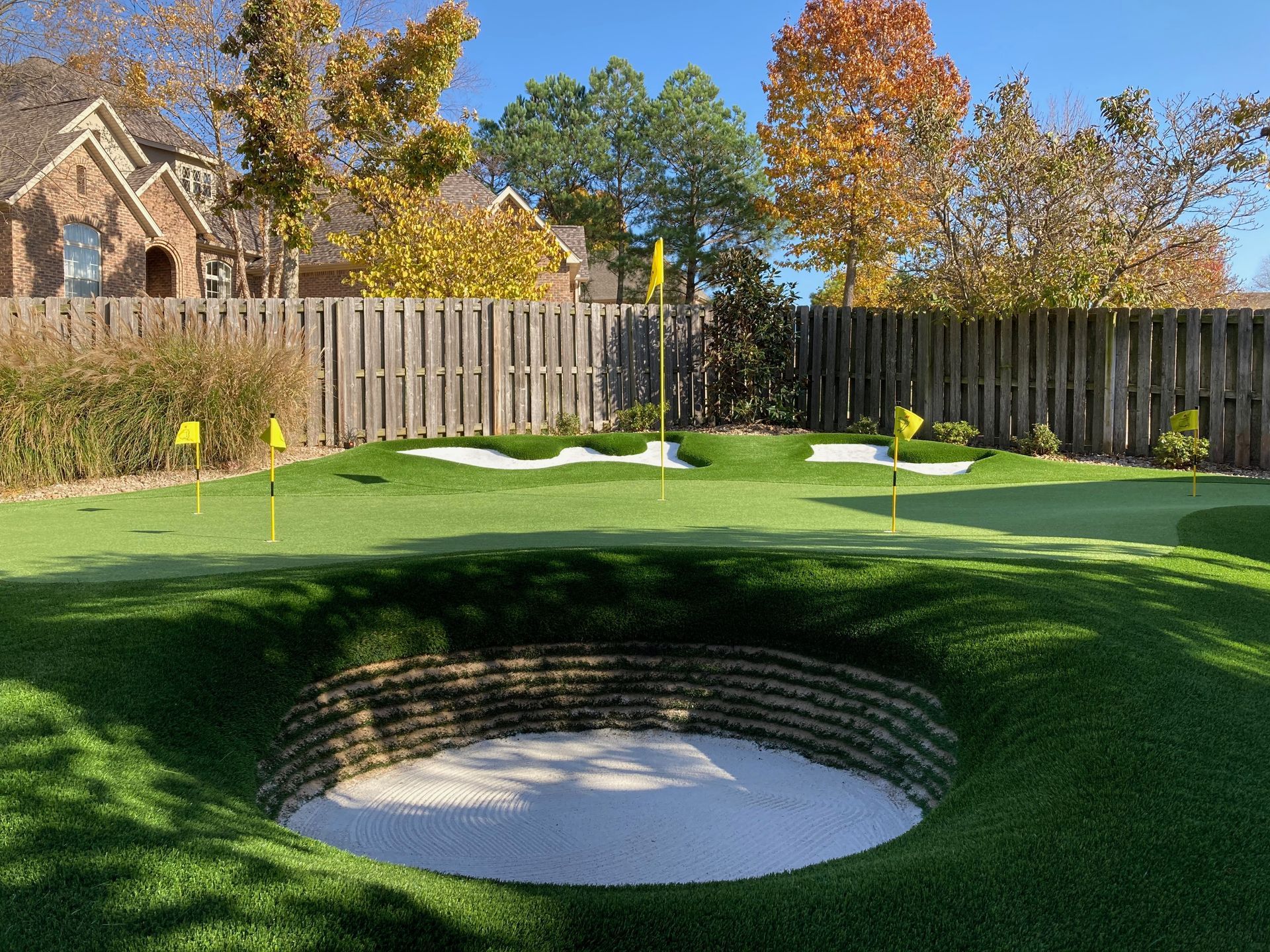 Golf green with a sand bunker, flagstick, and wooden fence under autumn trees