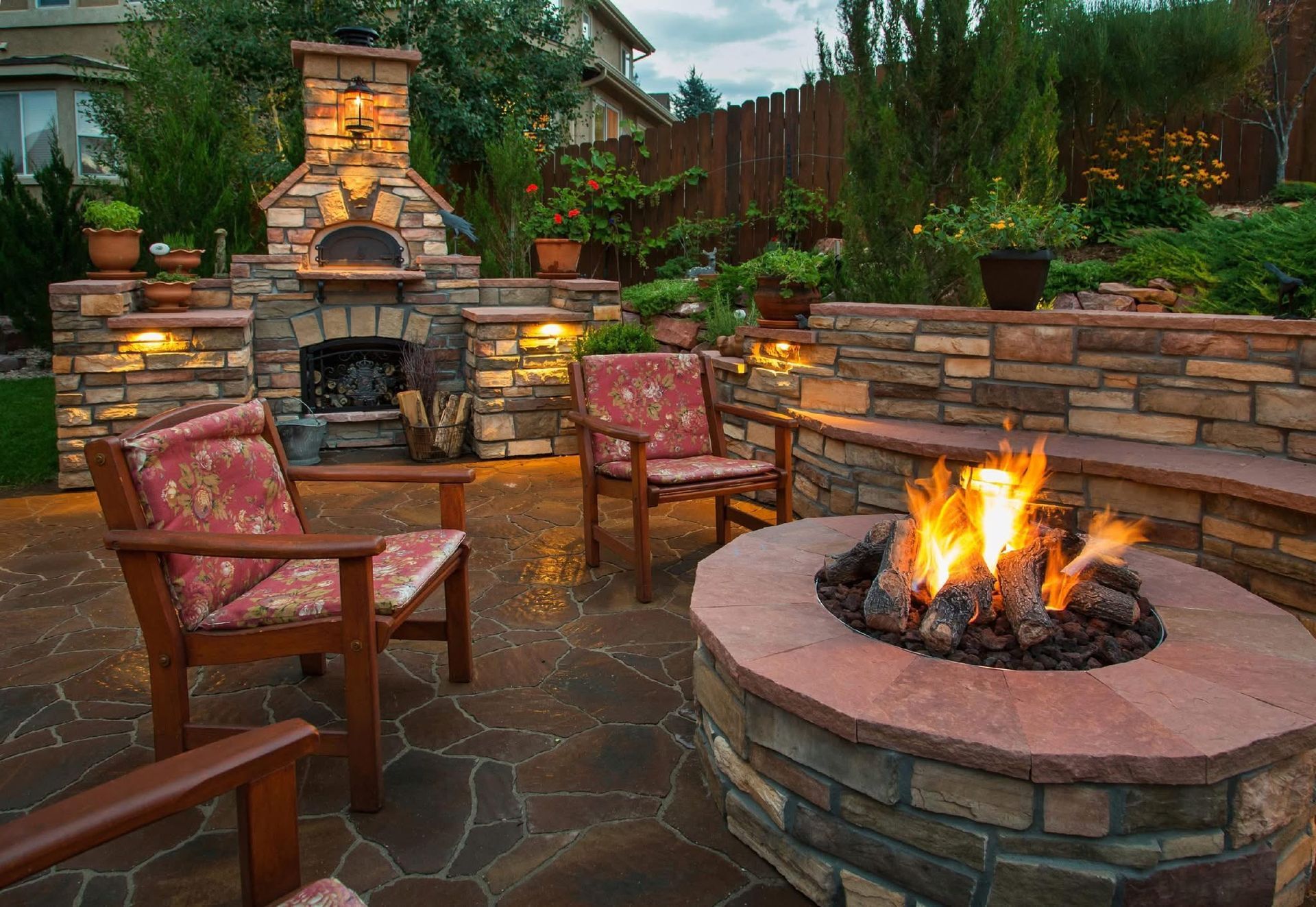Patio with stone fireplace, two red chairs, and a lit fire pit at dusk