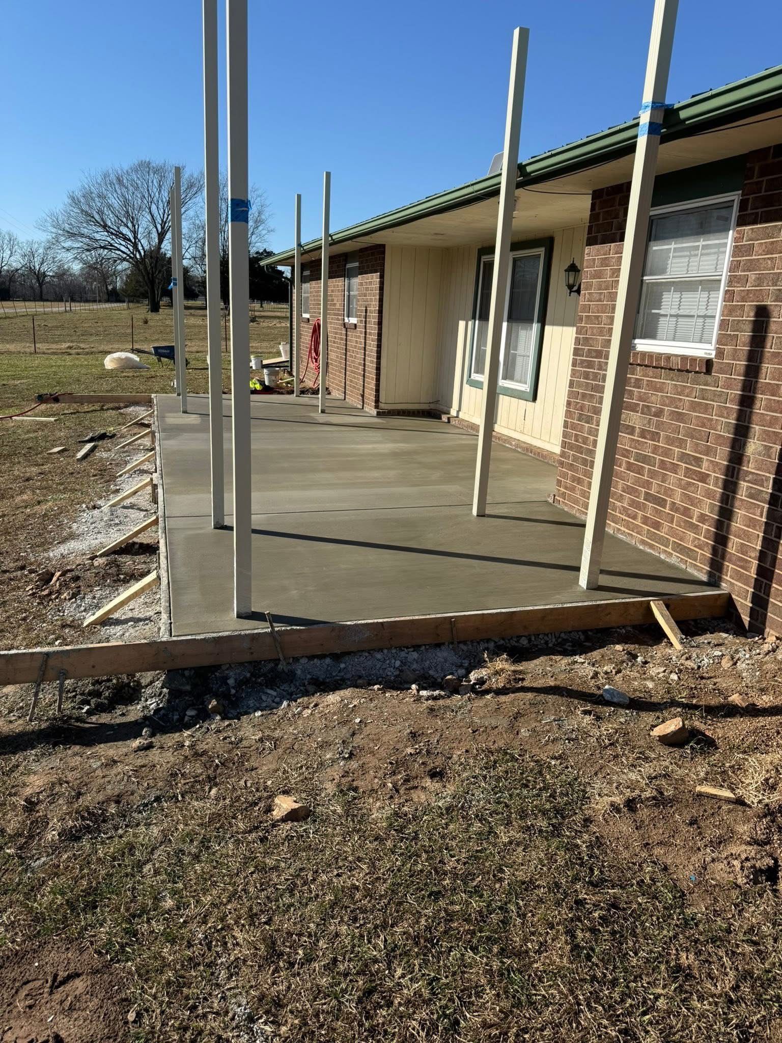 Unfinished concrete porch beside a brick house, with metal posts and gravel yard.