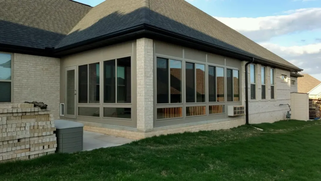 Suburban brick house with large sunroom windows and a sloped dark roof beside a grassy yard