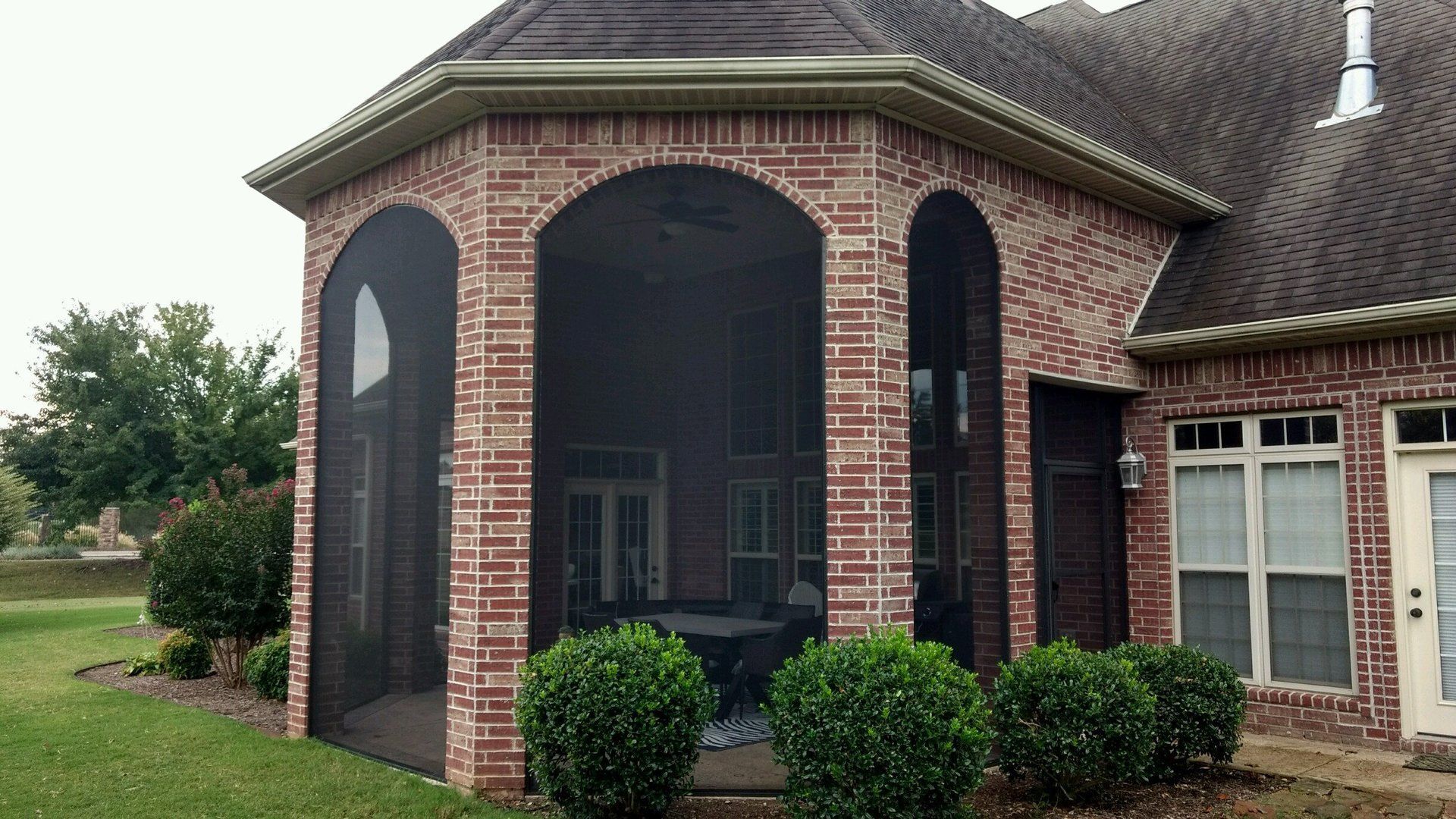 Brick house front porch with arched entry and dark roof, bordered by trimmed bushes