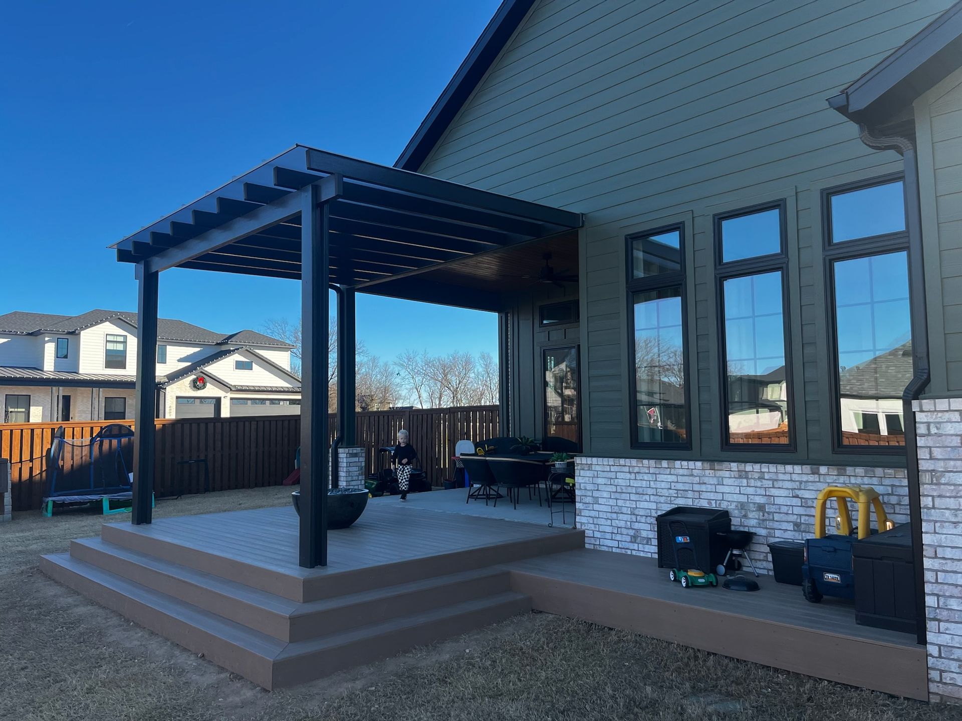 Back patio with gray pergola, steps, outdoor seating, and house siding in bright sunlight