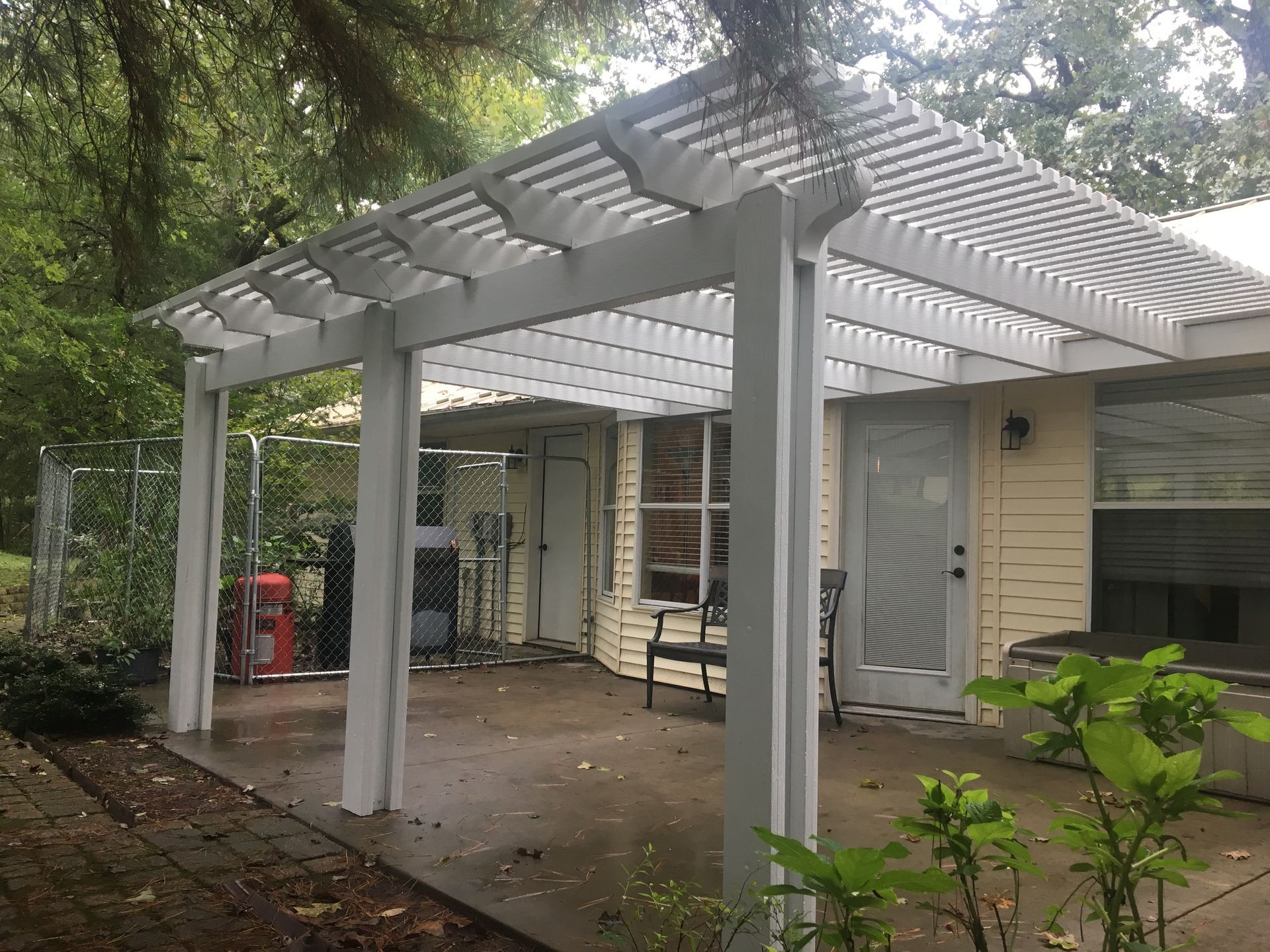White pergola-covered patio beside a house, with a bench, column posts, and trees overhead
