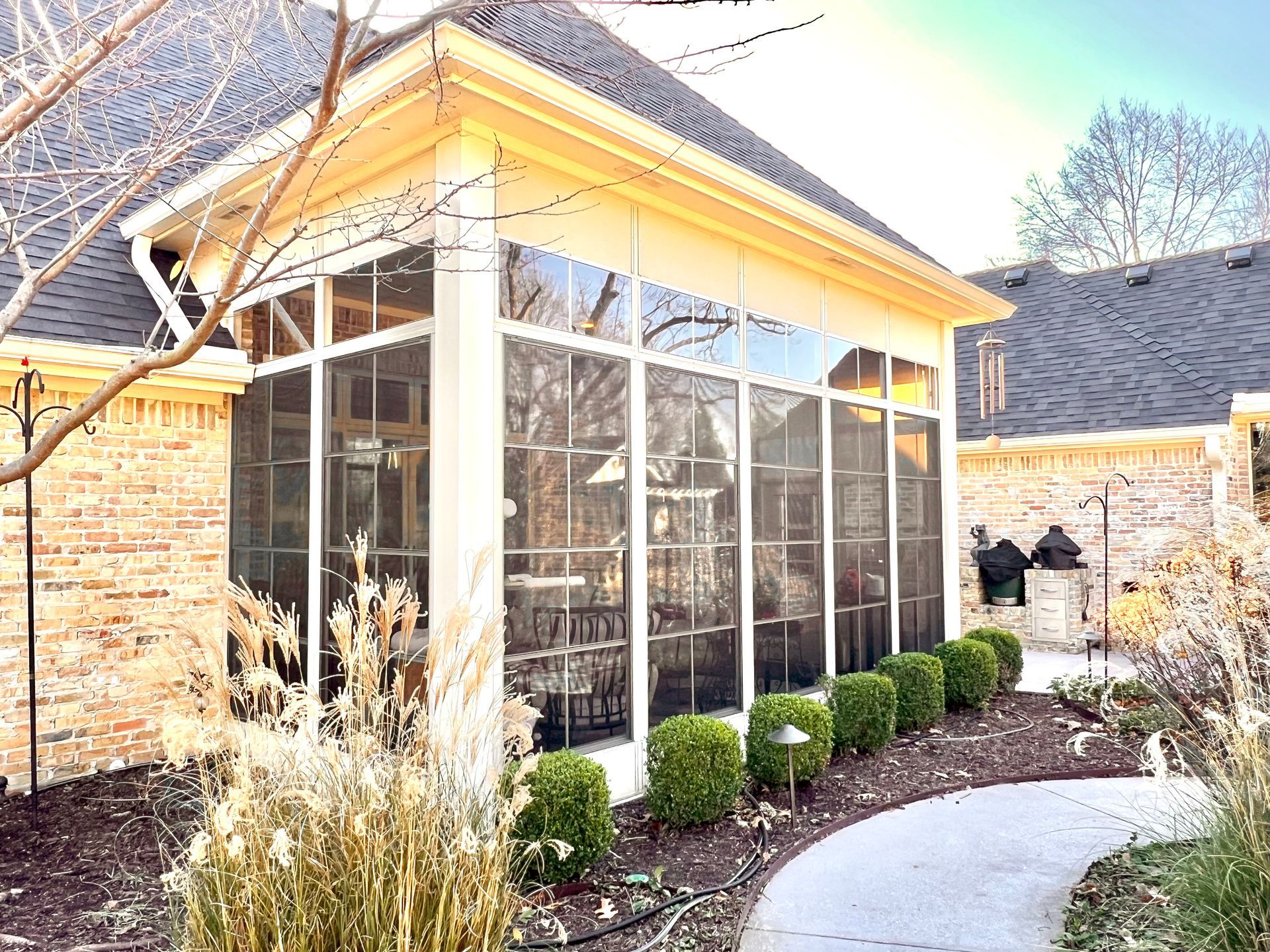 Sunlit glass sunroom beside a stone house, with potted shrubs and a curved walkway