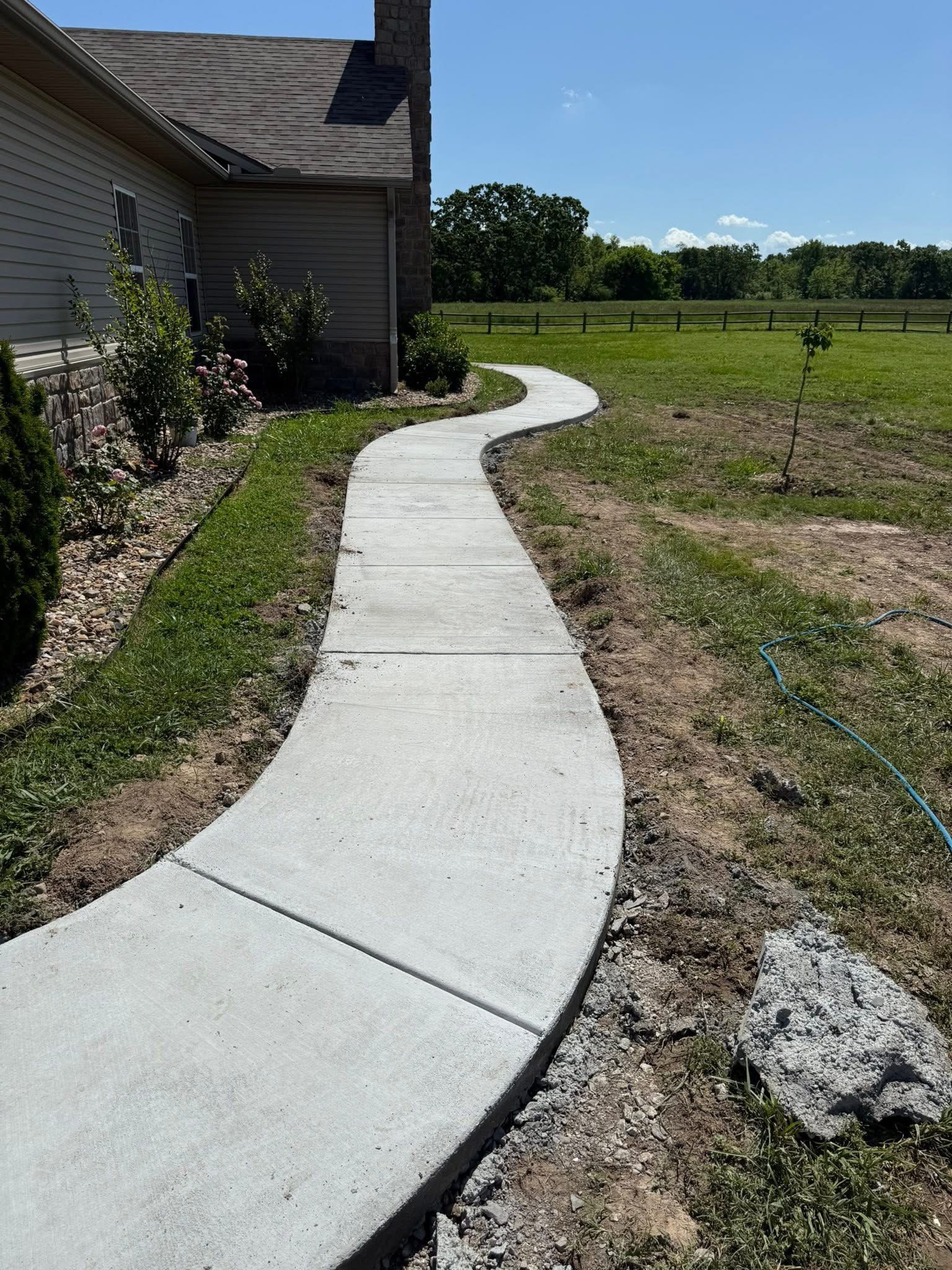 Curved concrete sidewalk beside a house, leading through a grassy yard toward open fields under a clear sky