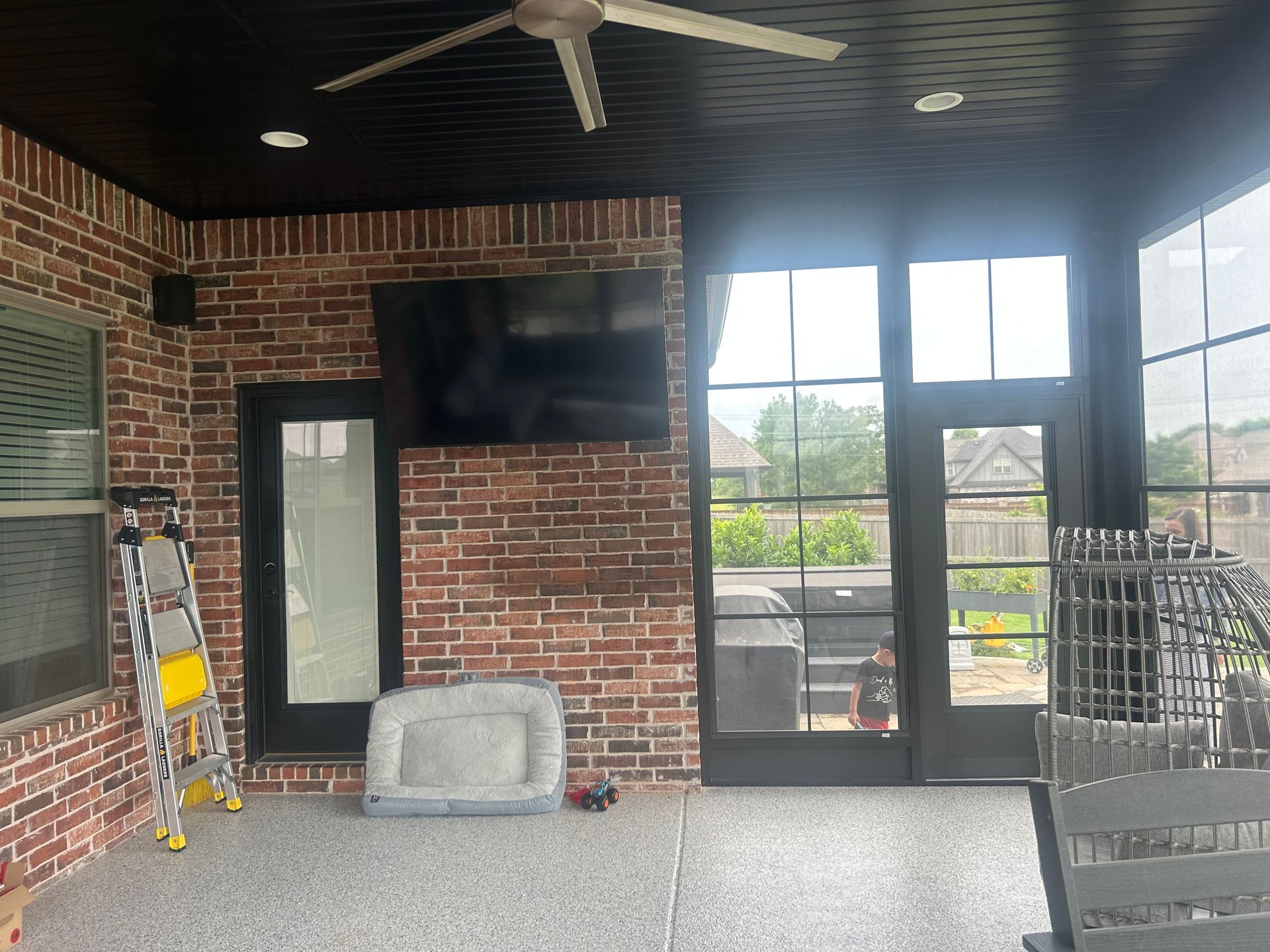 Covered brick patio with ceiling fan, TV, ladder, and large windows overlooking greenery.