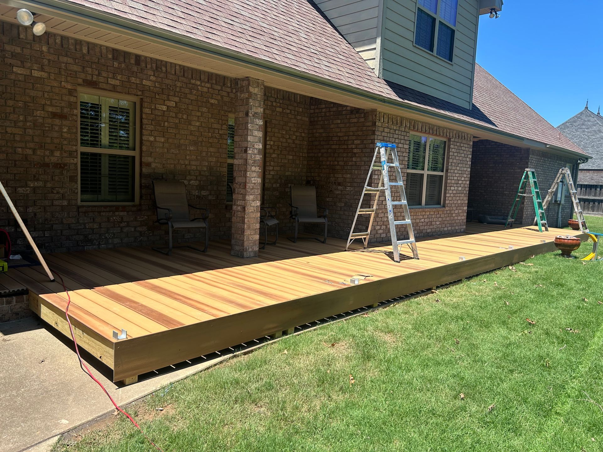 Wooden deck under construction beside a brick house, with ladders and tools on a sunny lawn.