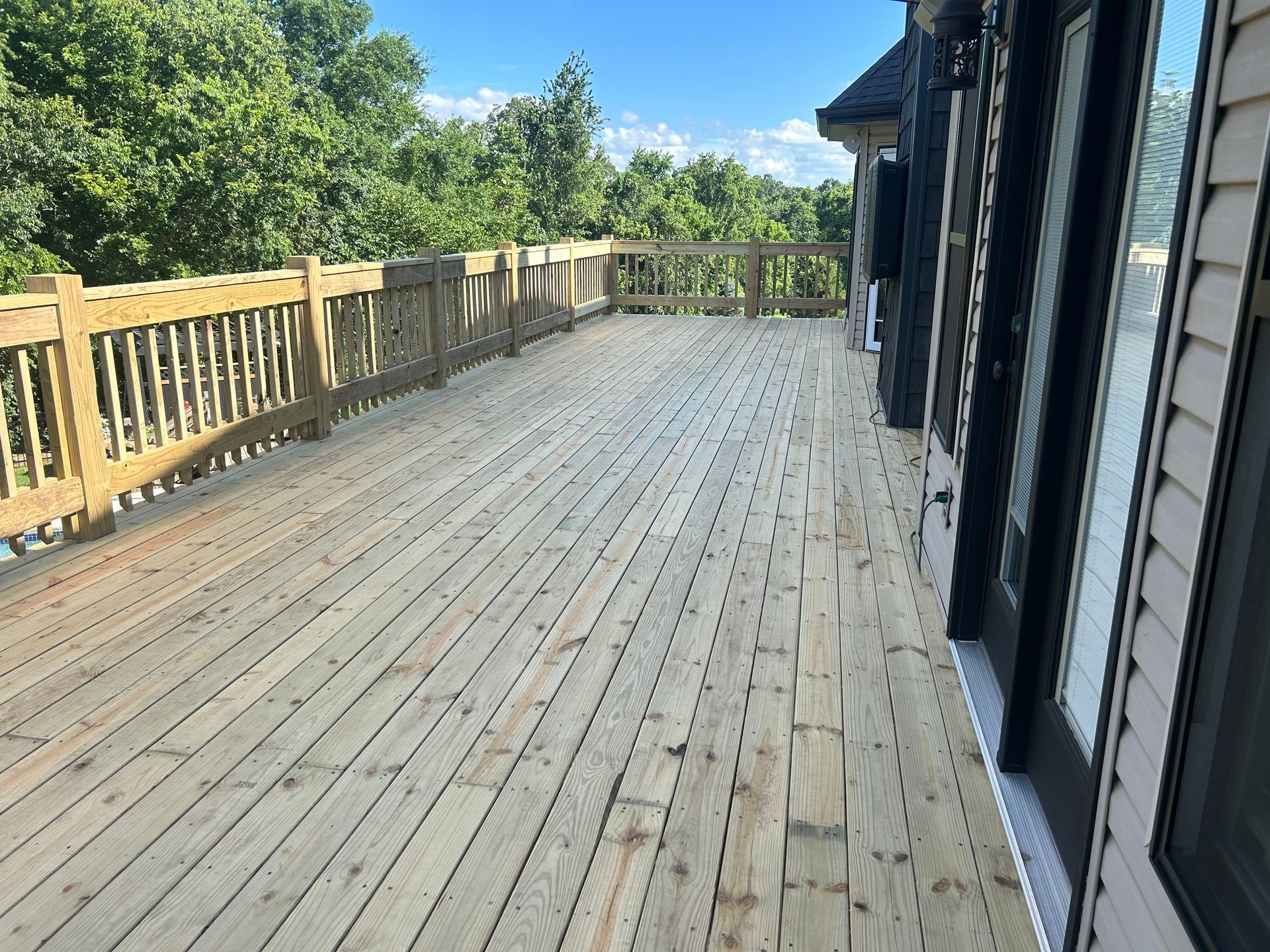Wooden deck beside a house, with railing and trees in bright daylight.