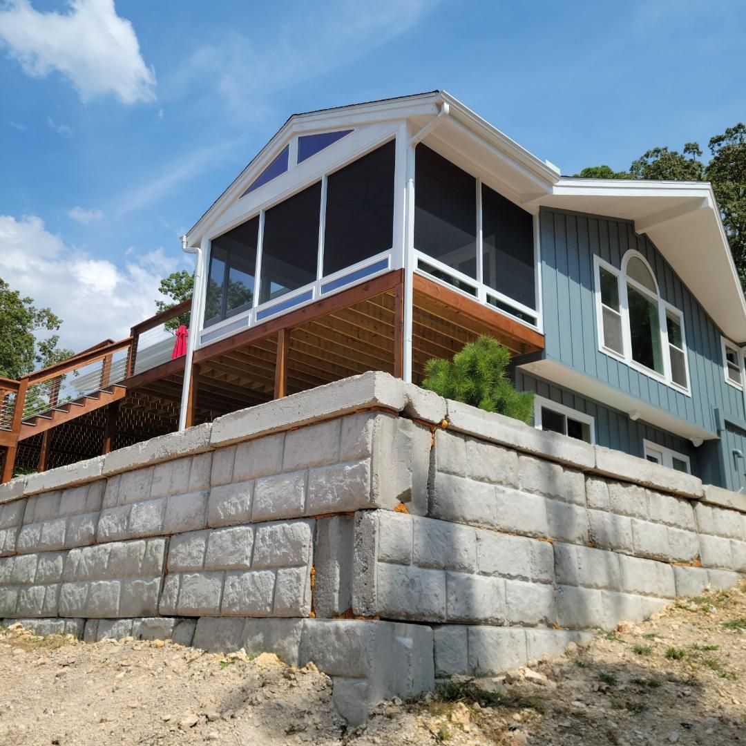 Blue house on a sloped lot with a screened porch and stone retaining wall under a sunny sky