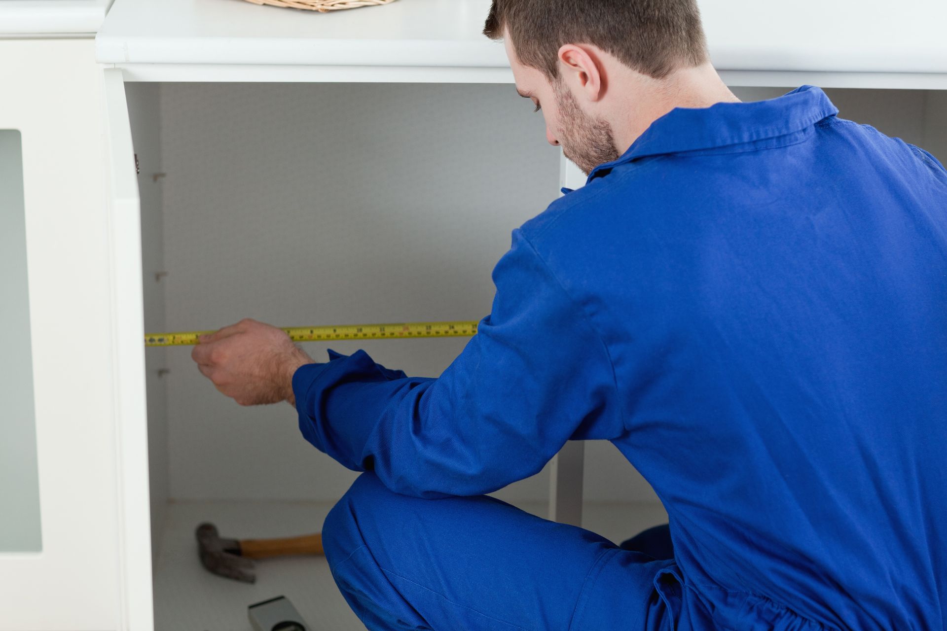 Man in blue measuring inside a white cabinet with a tape measure