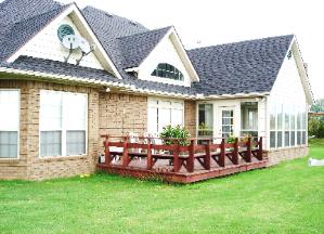 Brick house with a gray shingled roof, large windows, and a wooden deck on a green lawn