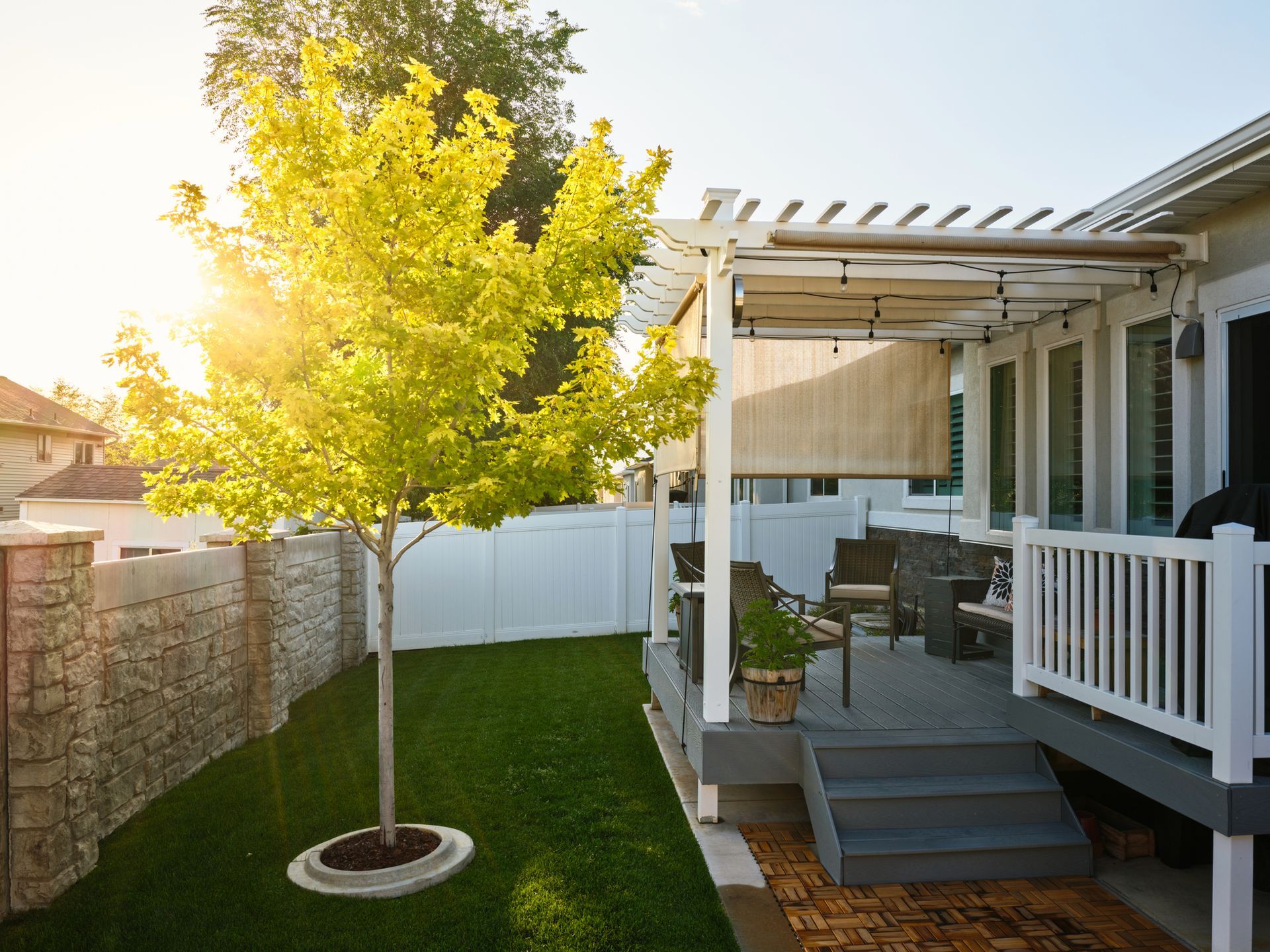 Sunlit backyard with a young tree, green lawn, white fence, and a small deck with patio furniture