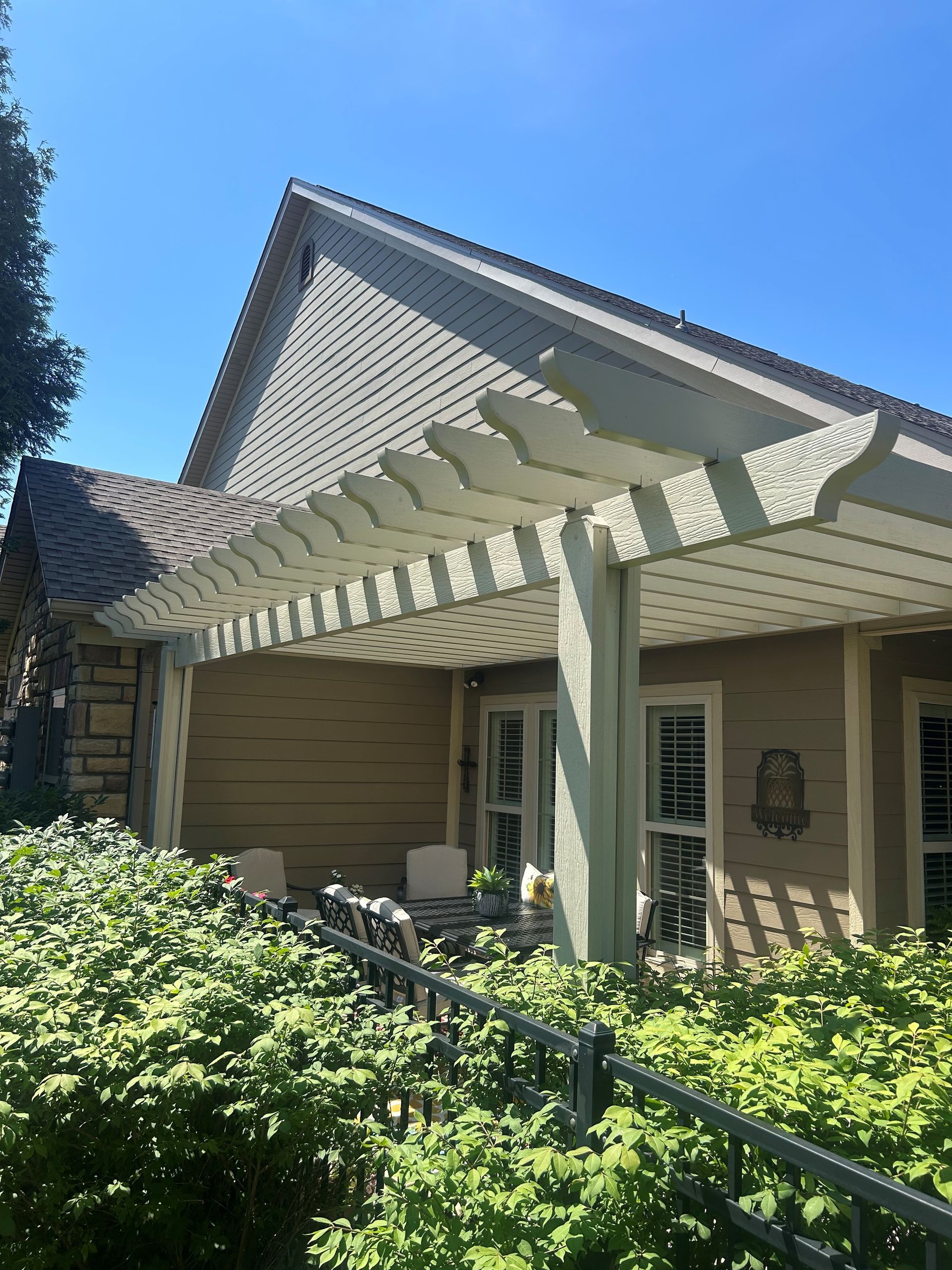 Covered porch with white beams and tan siding, framed by green shrubs under a blue sky.