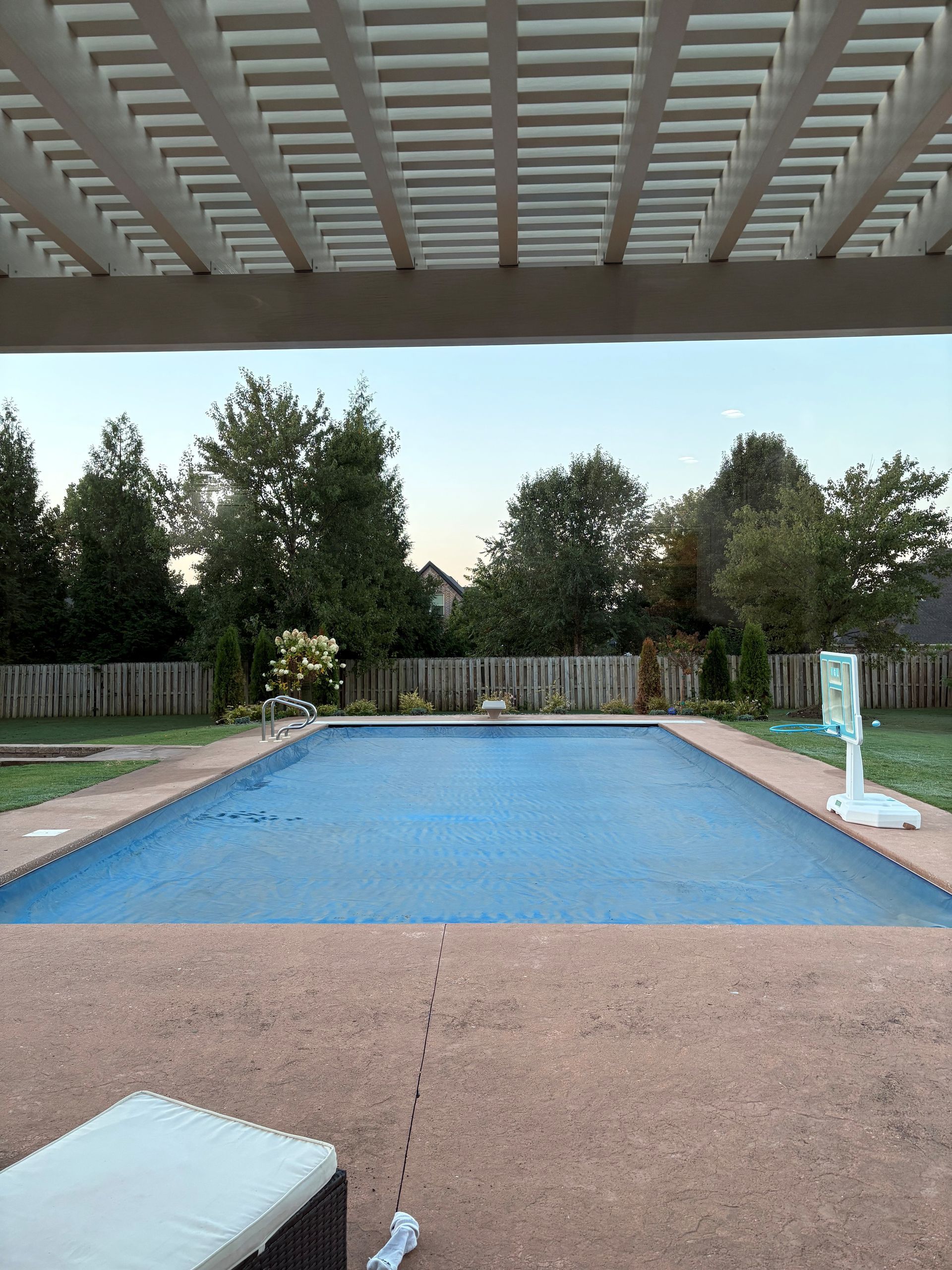 Outdoor backyard swimming pool under a covered patio, bordered by trees and a fence at dusk