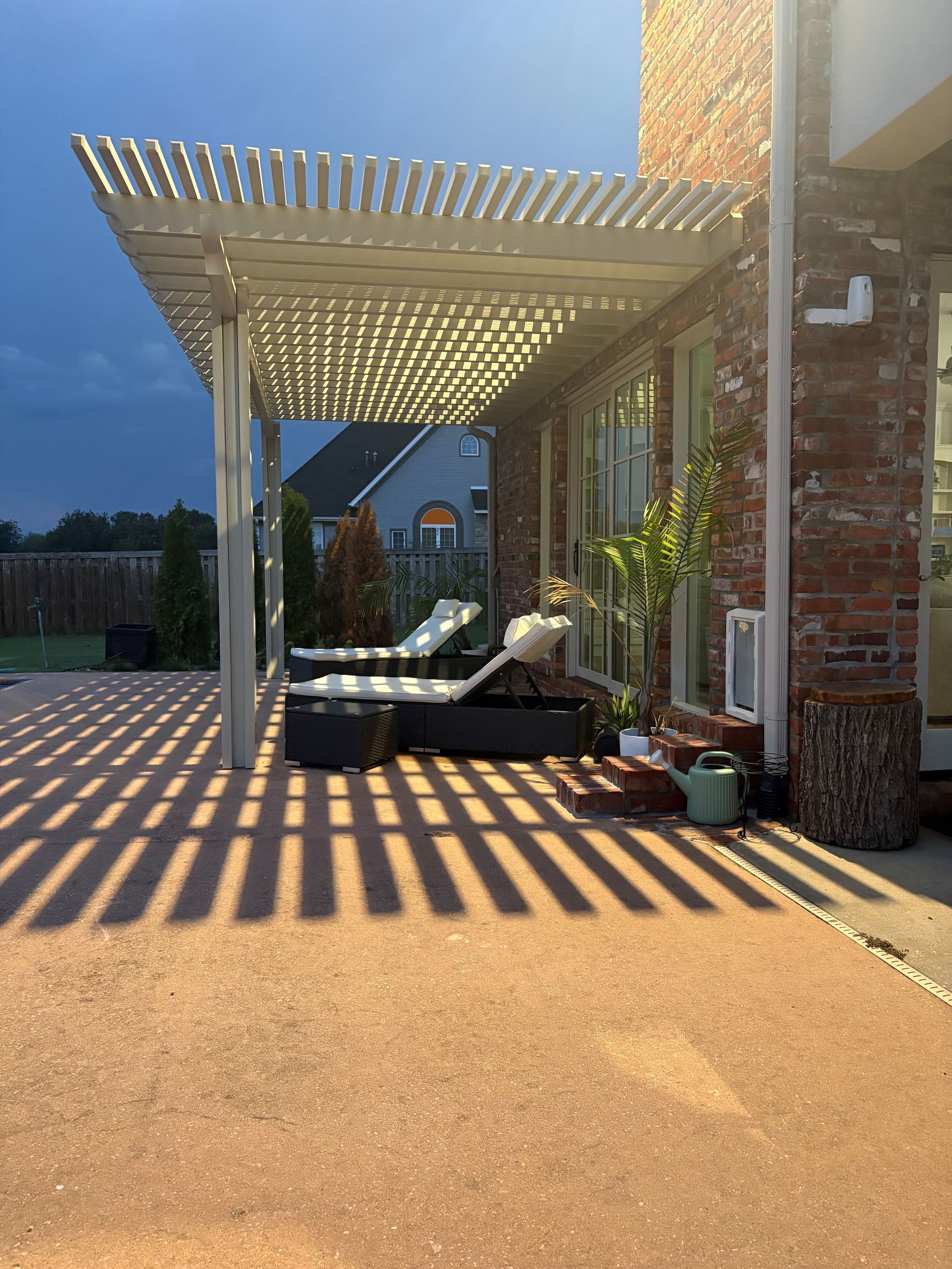 Patio with pergola casting striped shadows over outdoor seating beside a brick house at dusk
