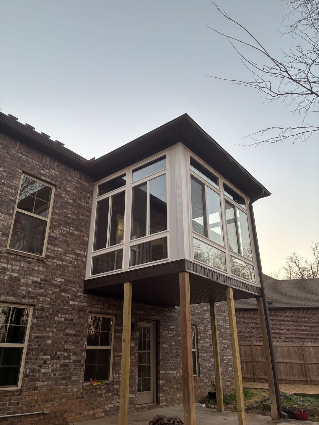 Two-story brick house with a raised glass sunroom on stilts and a small deck in a yard