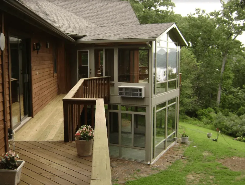 Wooden house deck with railing beside a glass-enclosed sunroom, overlooking a grassy hillside.