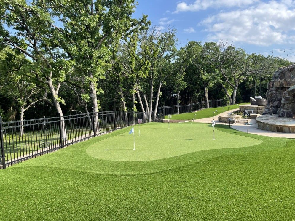Green mini-golf course with a putting green, fence, trees, and a rock waterfall feature under sunny skies