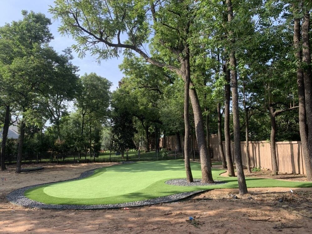 Tree-lined park with a small green putting area and wooden fence in the background