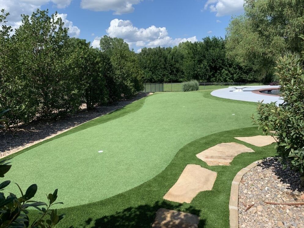 Backyard putting green with stone path, surrounded by trees and a pool on the right