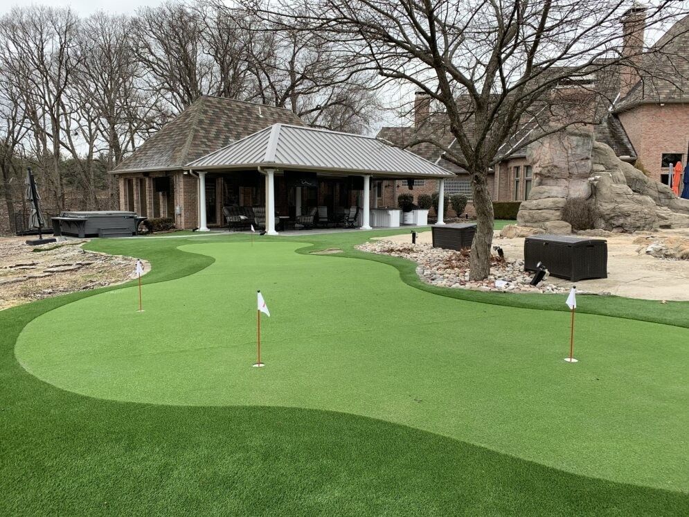 A putting green with flags beside a house and patio, bordered by trees and a stone landscaping feature.