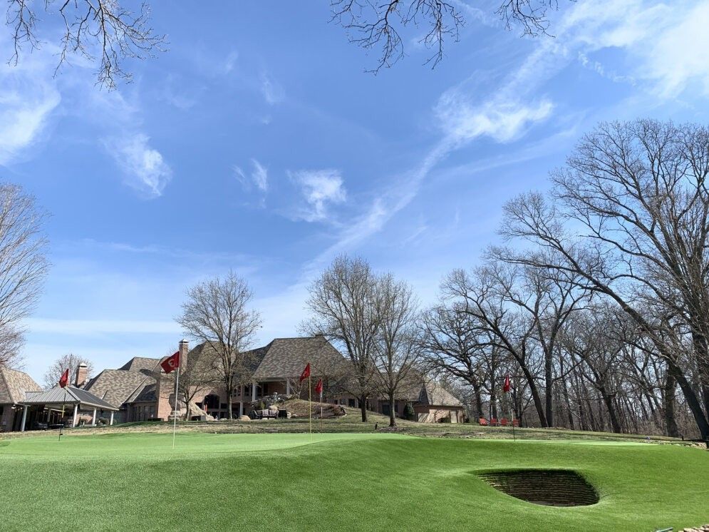 Golf course with green fairway, sand bunker, leafless trees, and houses under a blue sky