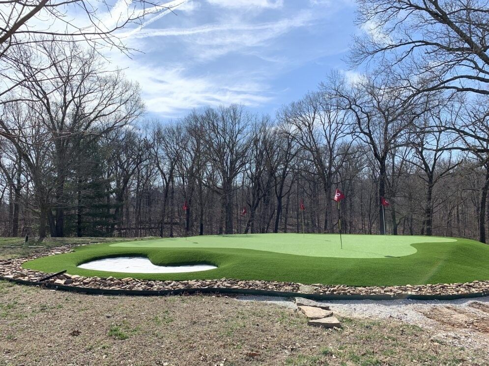 Golf green with a small sand bunker, surrounded by bare trees under a blue sky