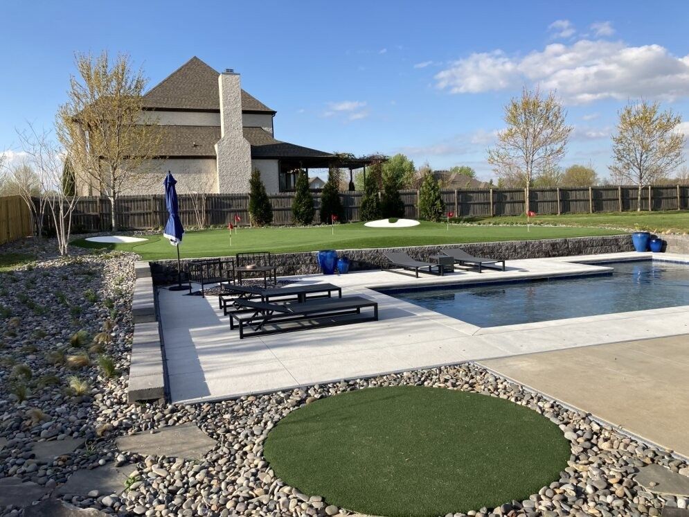 Backyard pool with lounge chairs, stone patio, and a house under a blue sky