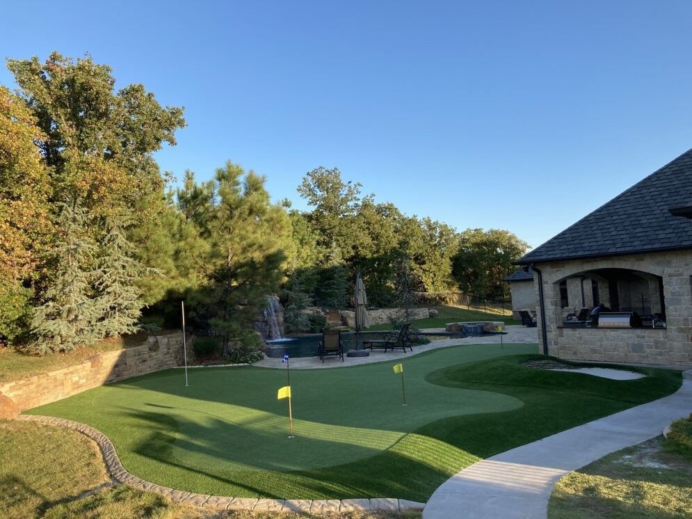 Backyard putting green beside a stone patio and house, with trees and a clear blue sky