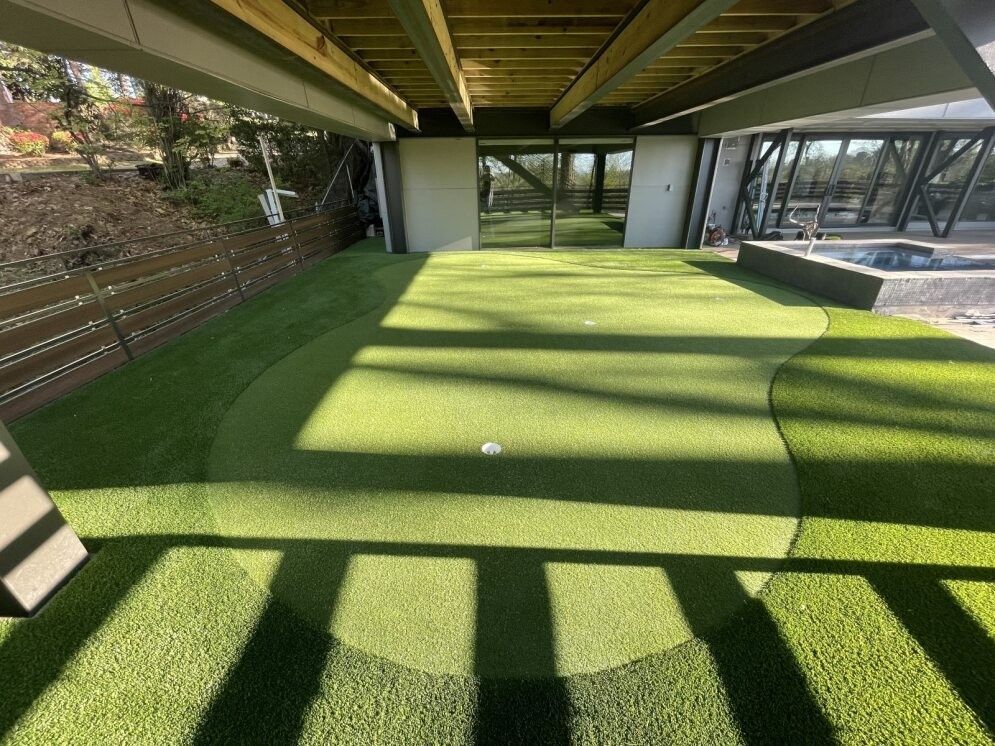 Shaded turf under a bridge with sunlit grass and concrete supports, beside a fenced walkway