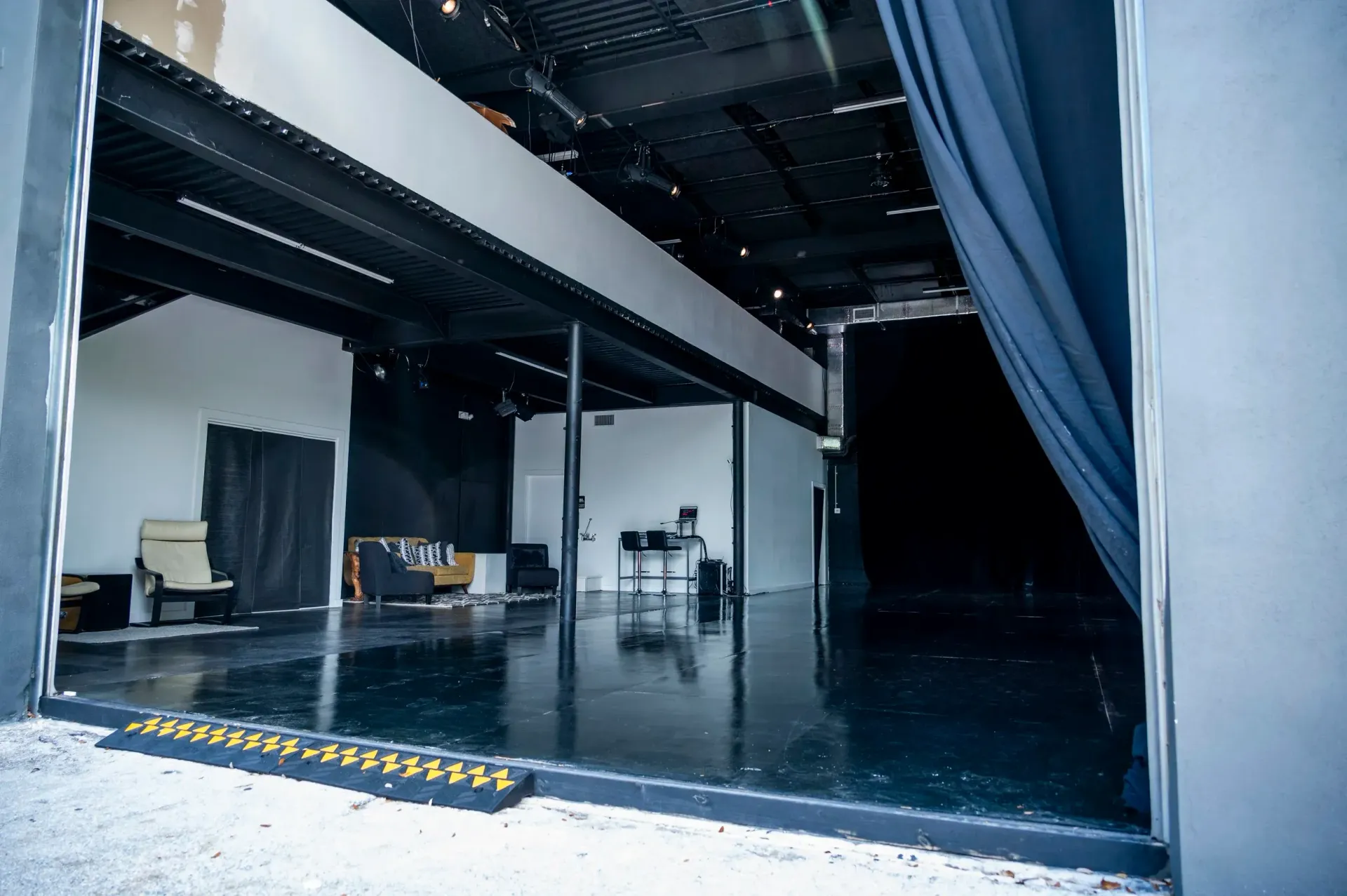 View into a black-walled theater space from a doorway, with some chairs, boxes, and a piano.