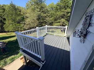 A dark-colored wooden deck with a white railing overlooks a grassy yard and dense green trees under a clear blue sky.