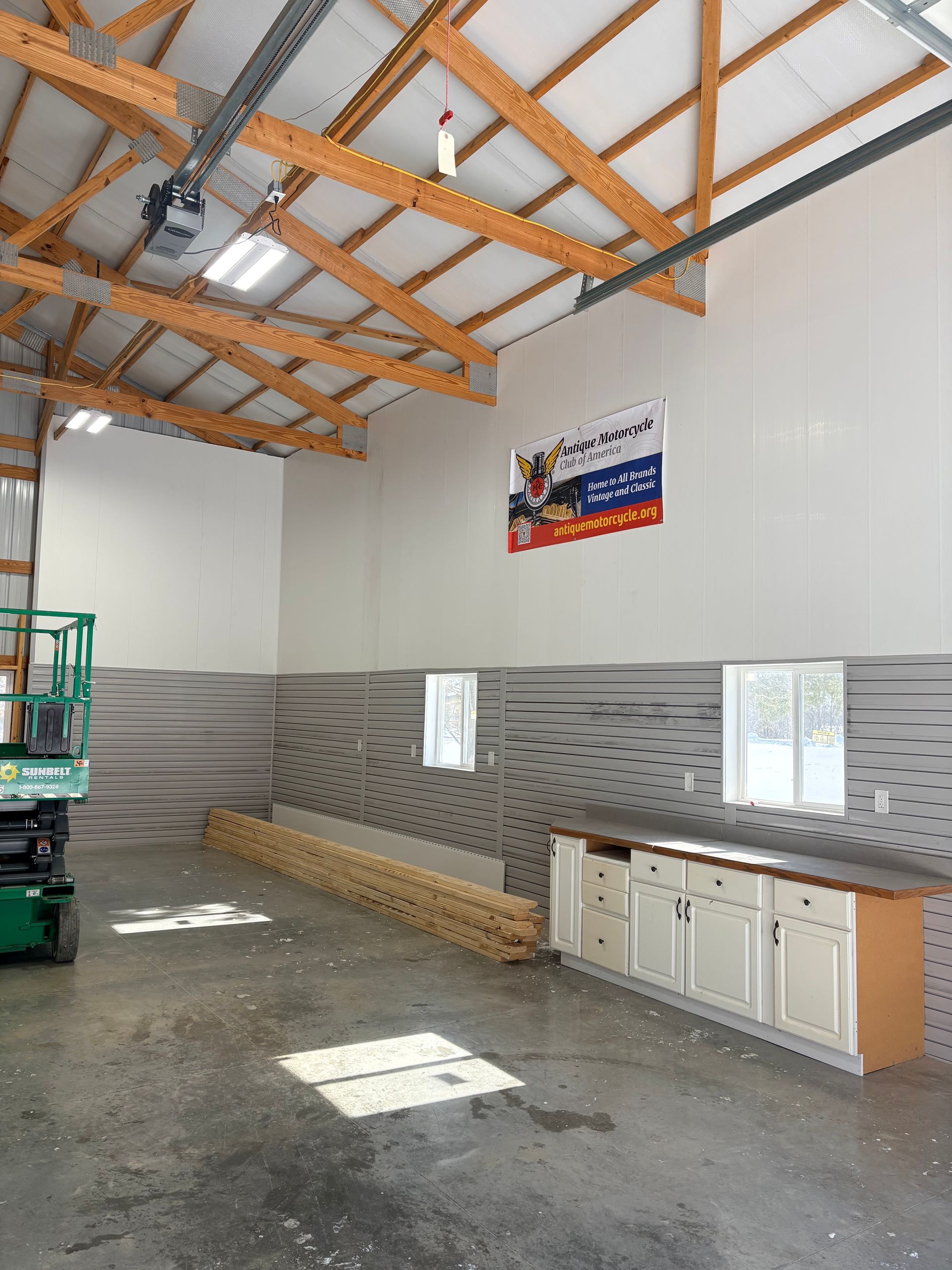 Large industrial workshop with a wooden ceiling truss, gray horizontal-striped wall panels, a work counter, and a scissor lift.