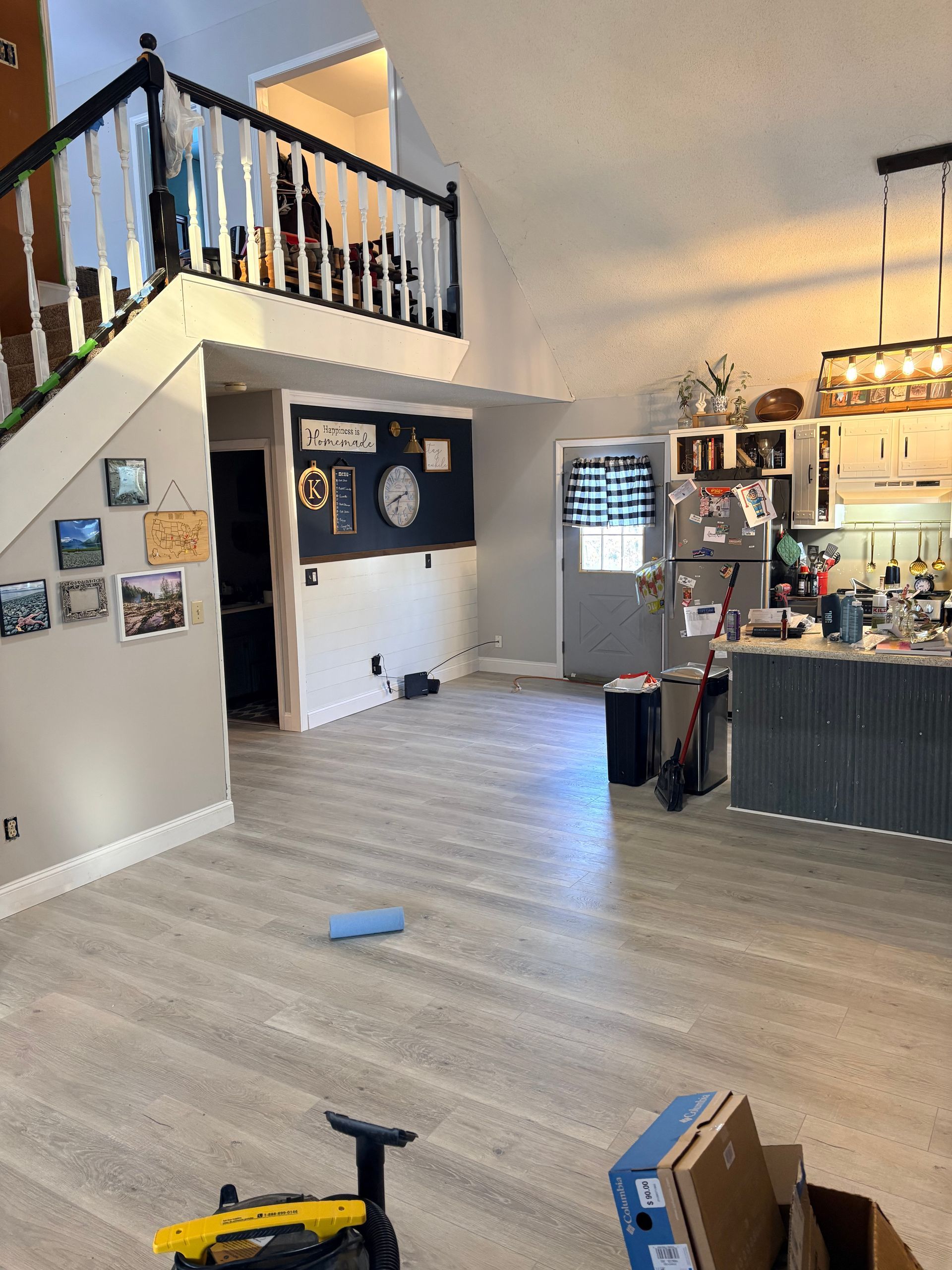 An open-plan home interior featuring light wood flooring, a staircase with a black railing, and a kitchen in the background.