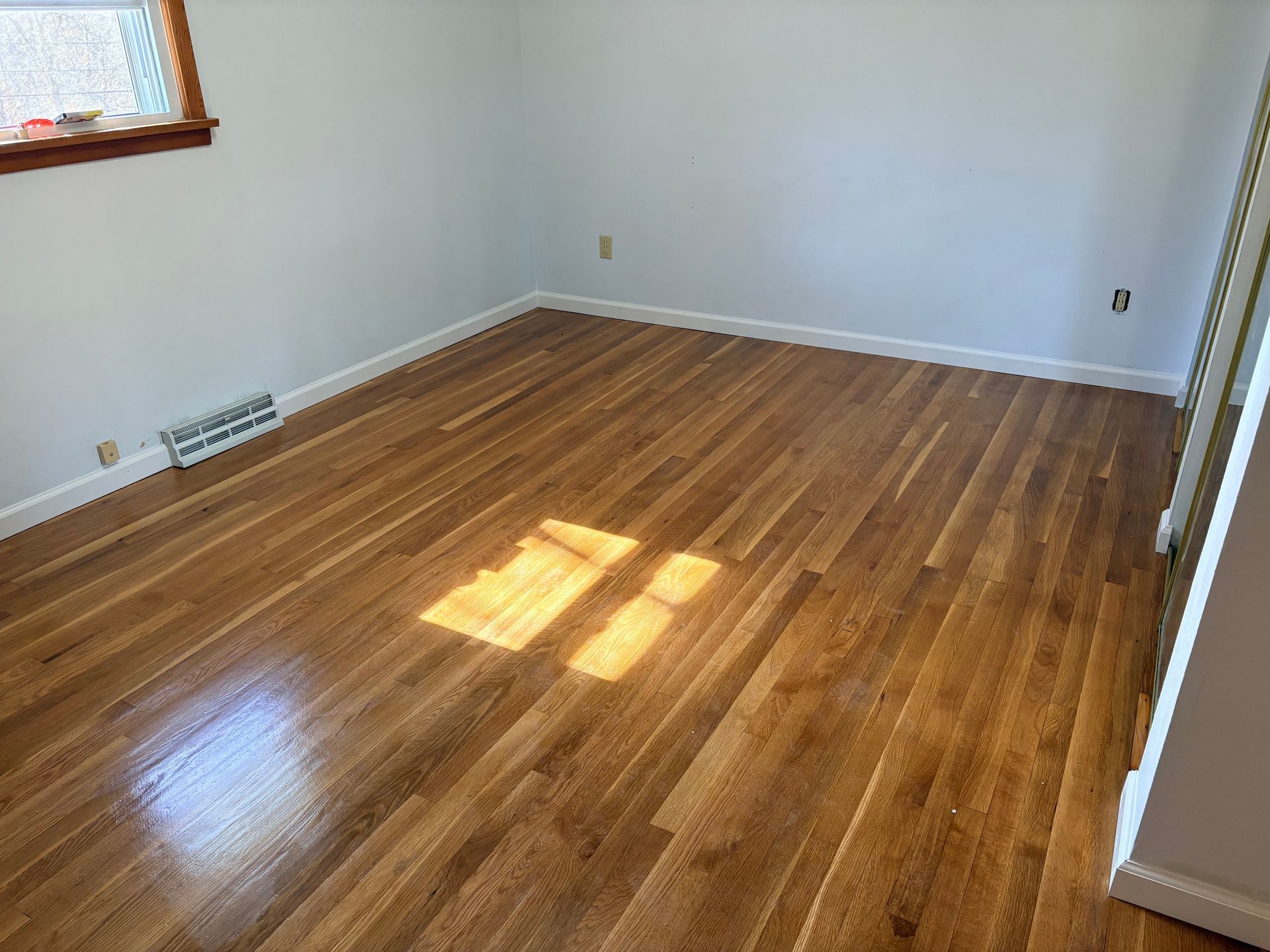 Empty room with polished light wood floors, grey walls, and a window reflecting sunlight onto the floor.