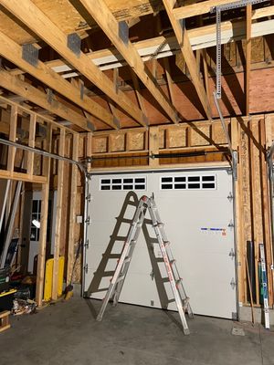 An open-framed garage interior under construction with a white sectional garage door and a metal stepladder in front.
