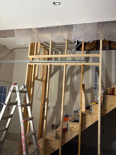 A wooden wall frame under construction in front of an unfinished staircase, with a ladder in the foreground.