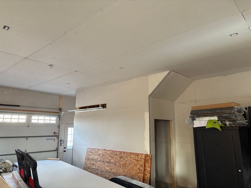 An unfinished garage interior with exposed drywall, ceiling panels, and a staircase alcove leading to a doorway.
