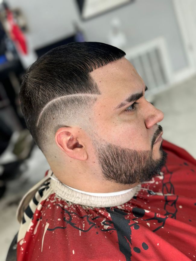 Side profile of a man with a fresh, detailed skin fade haircut and a sculpted beard, wearing a red patterned barber cape.