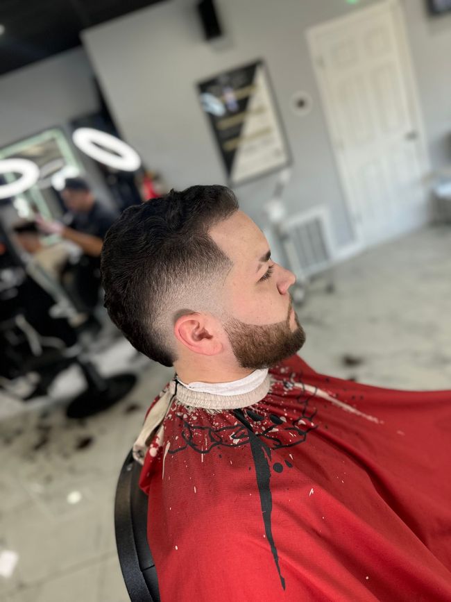A person with a fresh fade haircut and beard trim sitting in a barbershop chair wearing a red salon cape.