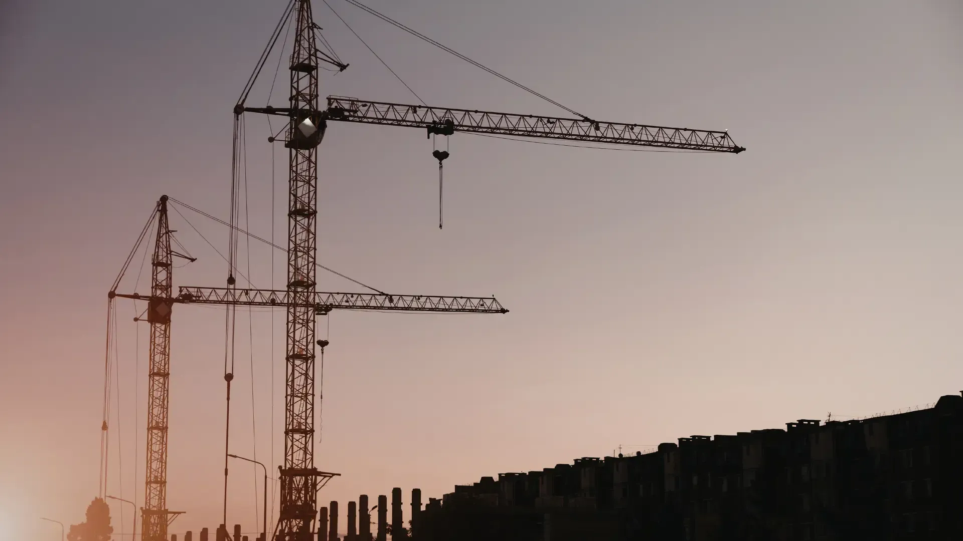 Two construction cranes silhouetted against a pink dusk sky near a dark building rooftop