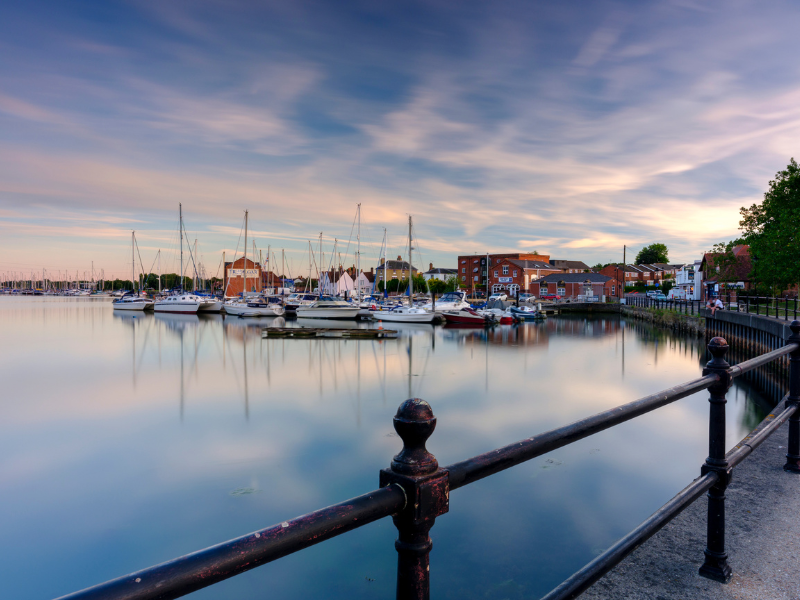 Marina with sailboats at sunset, calm water reflecting pastel sky and waterfront buildings