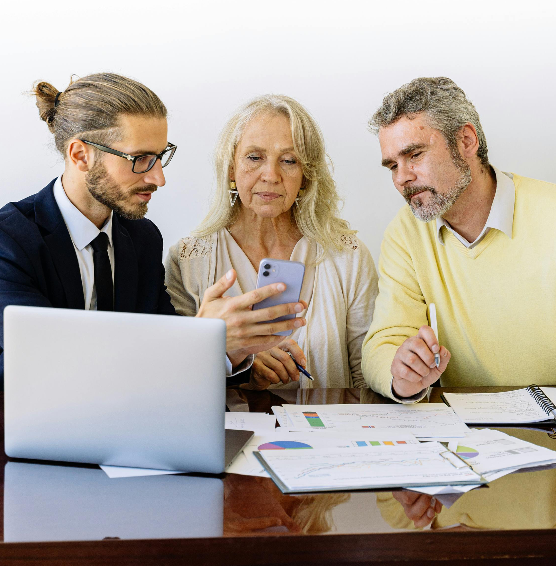 A professional meeting where three people review digital data on a phone, a laptop, and paper charts at a table.