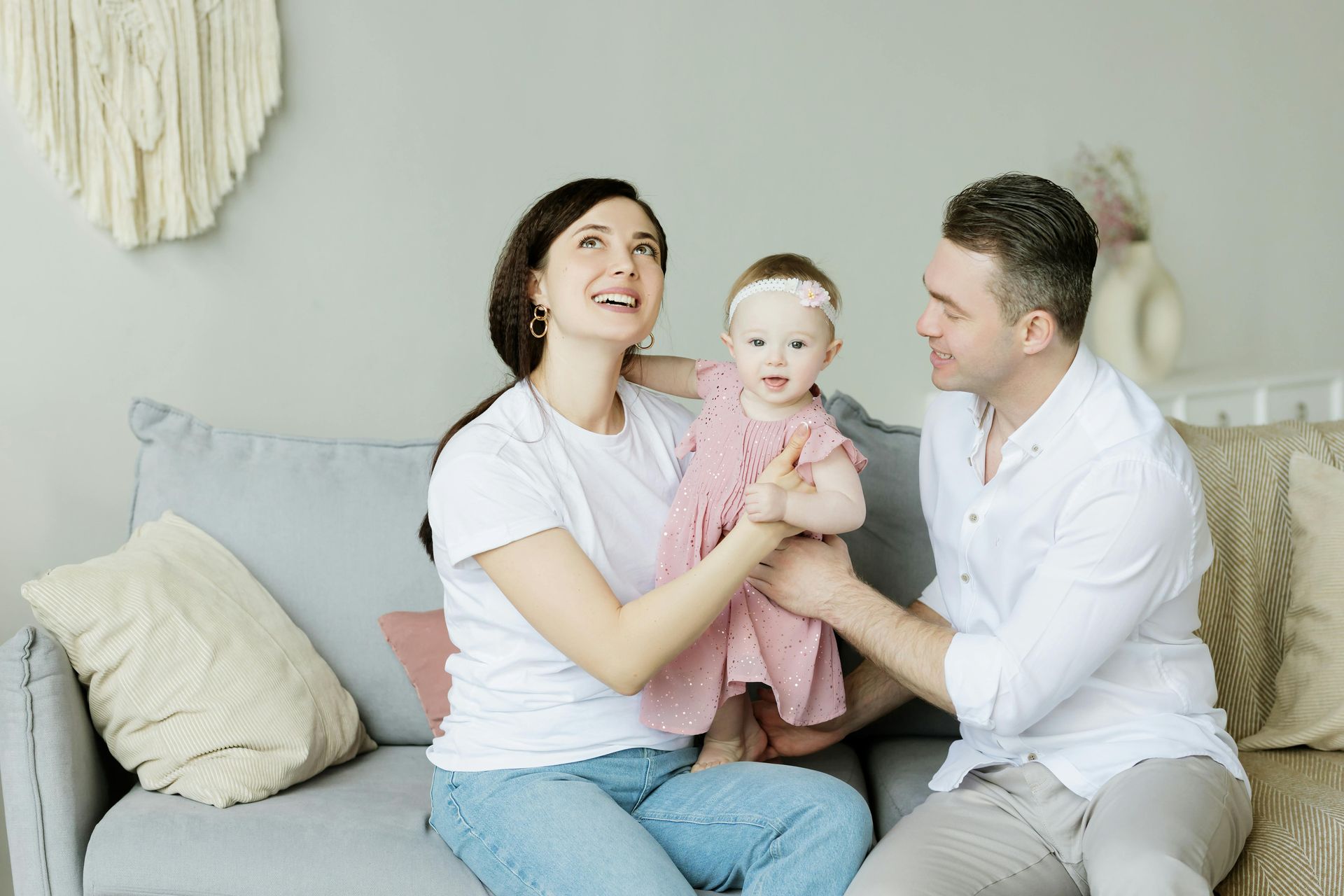 A small family sits together on a grey sofa in a bright room, looking toward each other and smiling.