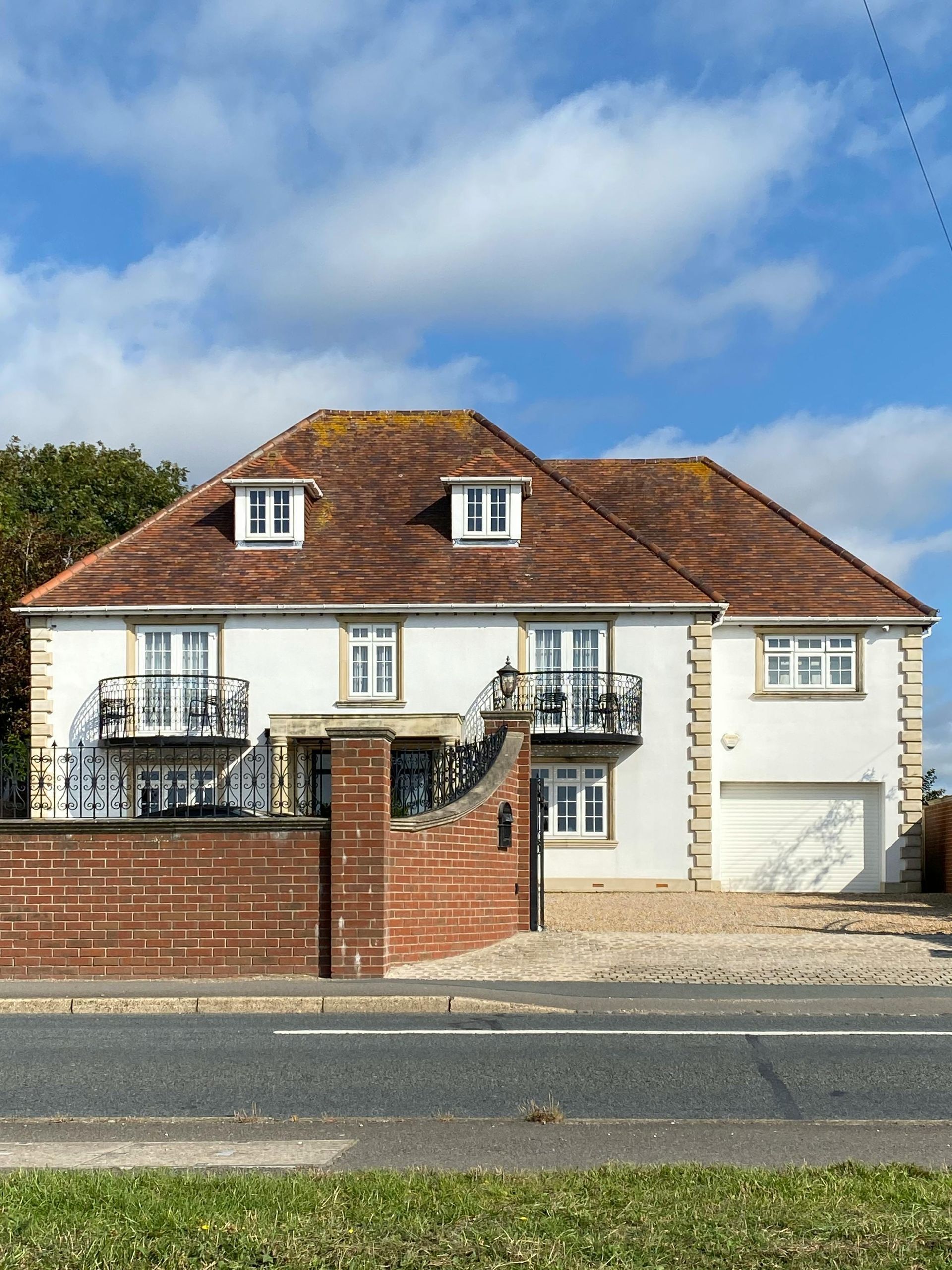 A two-story white house with a red tiled roof, balconies, and an integrated garage, behind a brick wall under a blue sky.